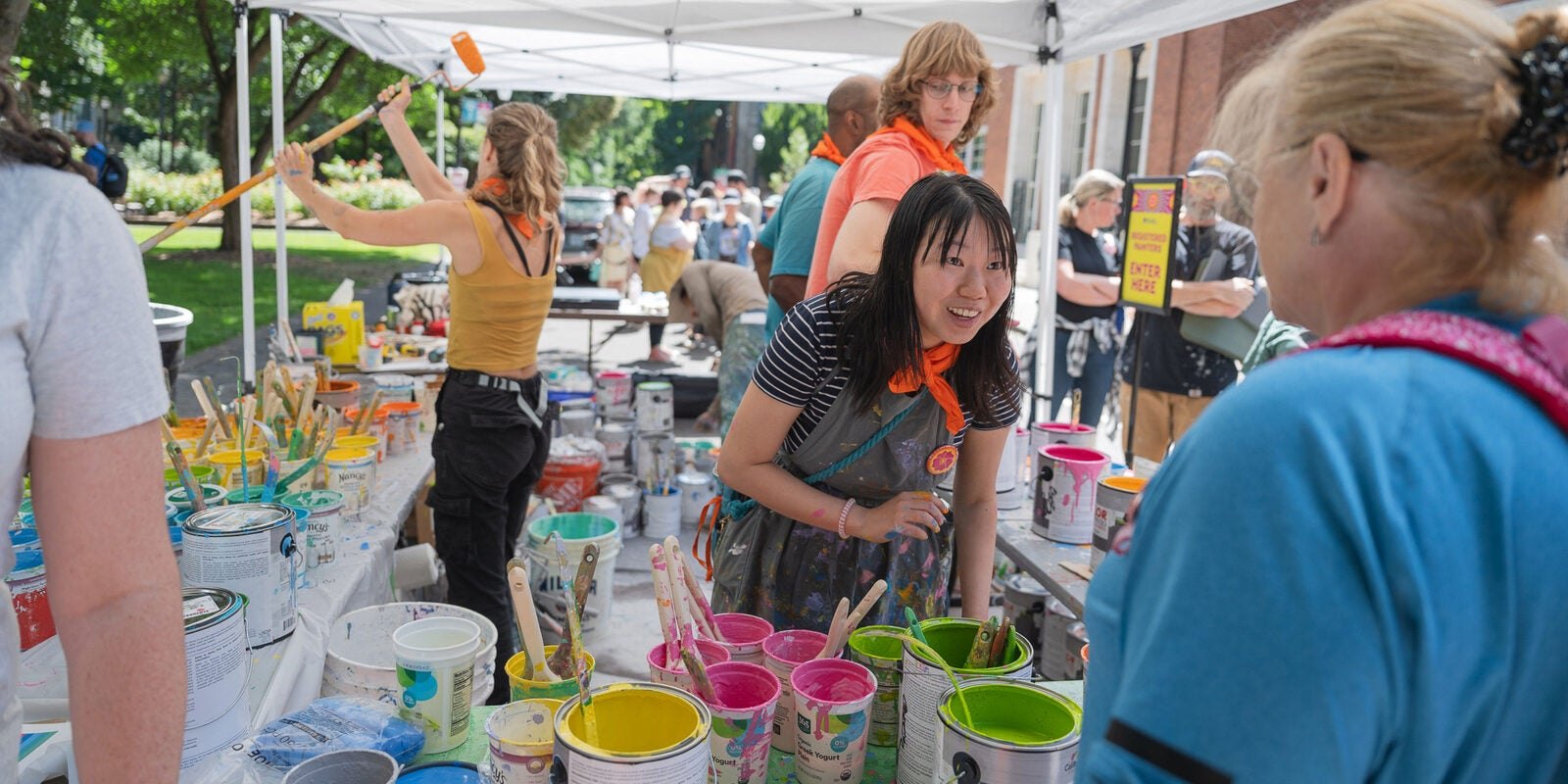 People stand under a white tent around tables covered with cans of colorful paint.