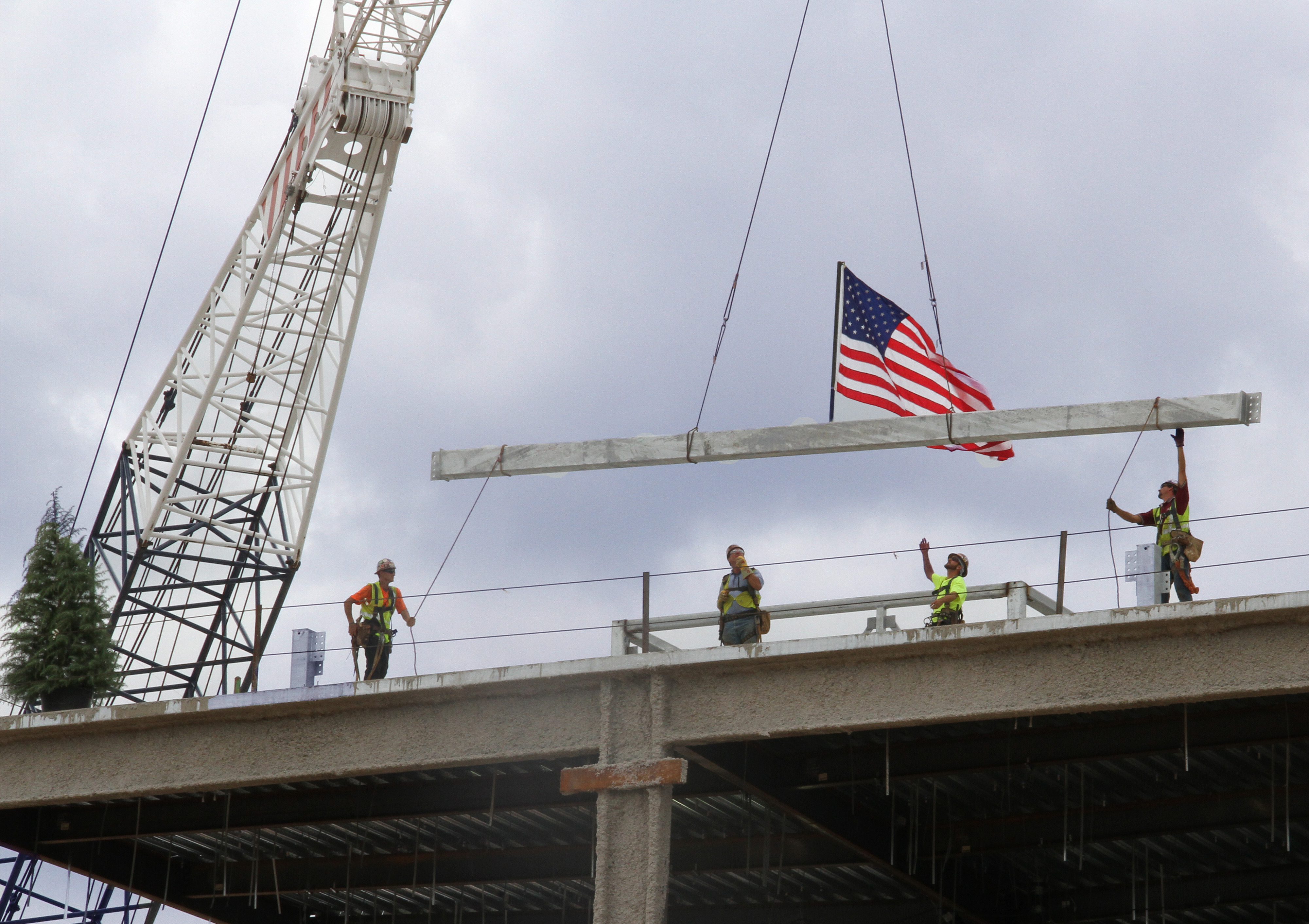 Workers in safety vests on a rooftop guide a large steel beam bearing an American flag lowered by a crane.