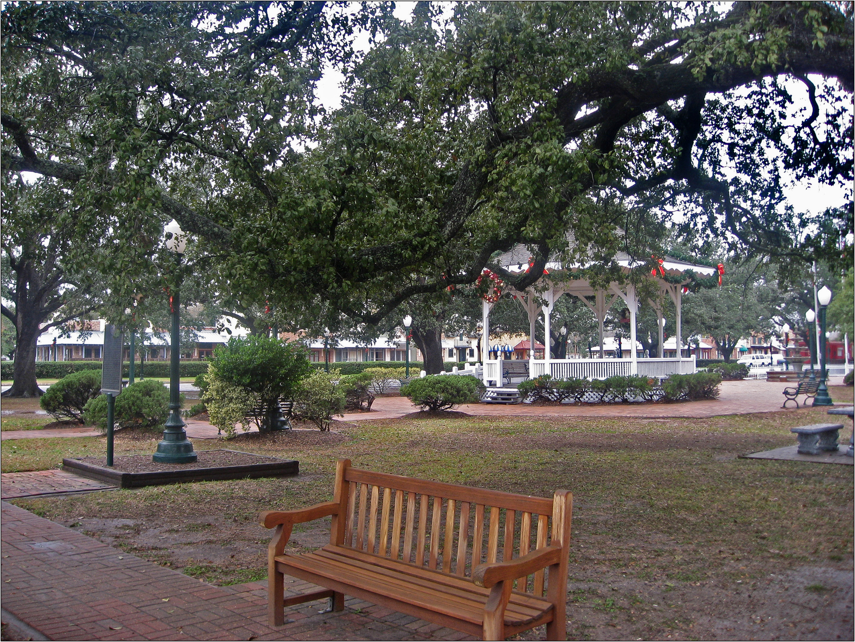 A white gazebo and wooden bench beneath large oak trees in a public park.