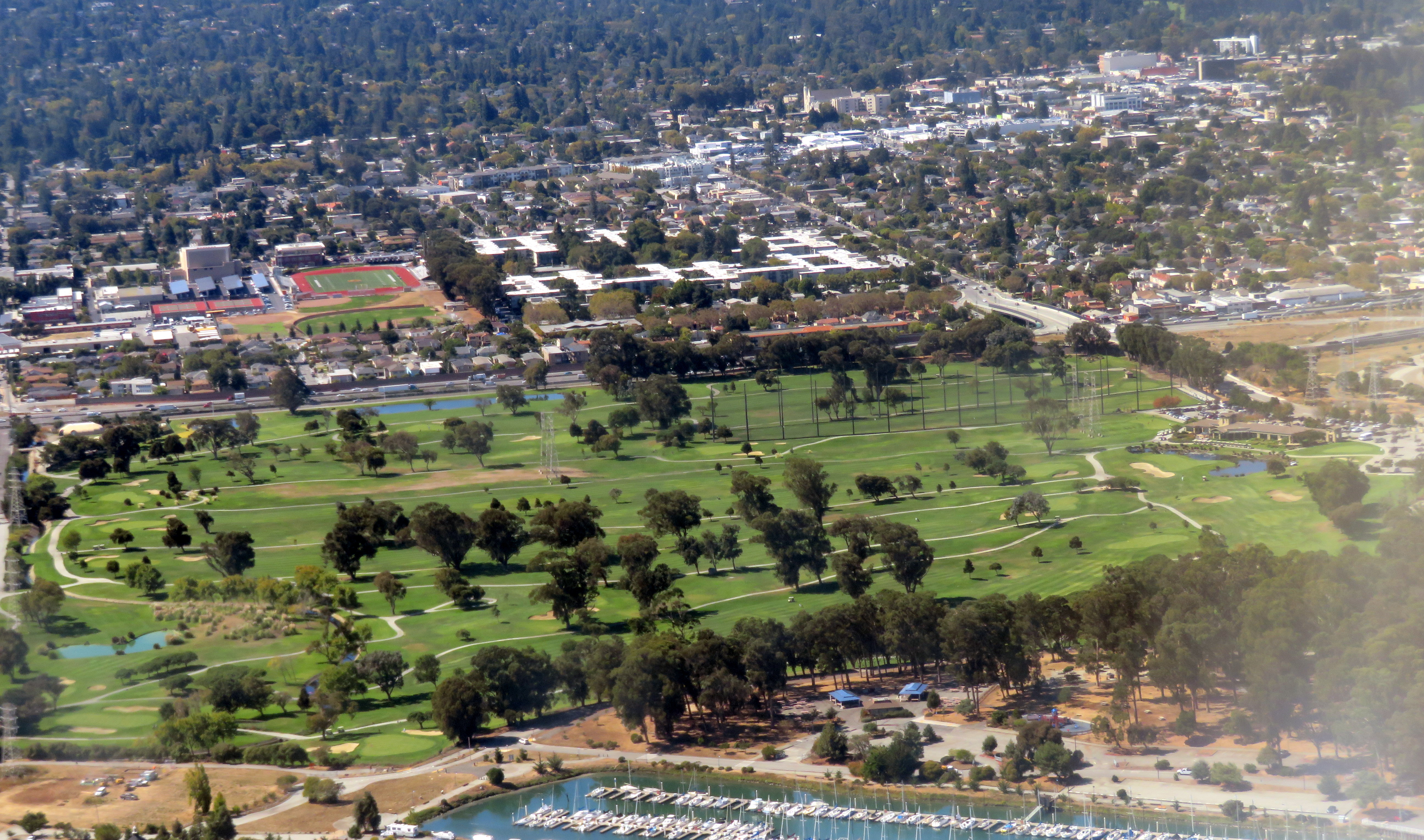 Aerial view of a golf course surrounded by trees with a marina and city in the background.