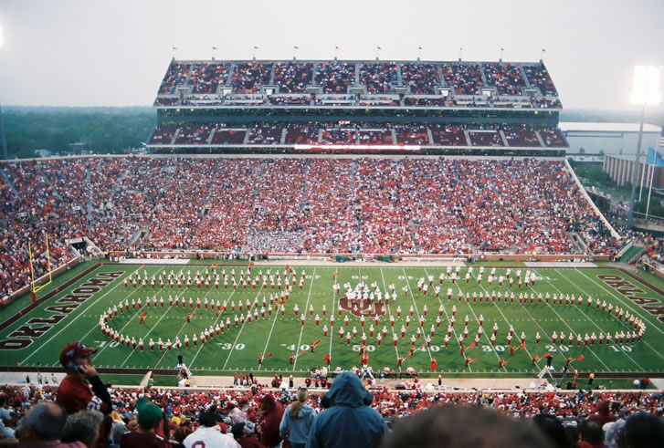 Packed college football stadium with a marching band performing on the field at halftime.