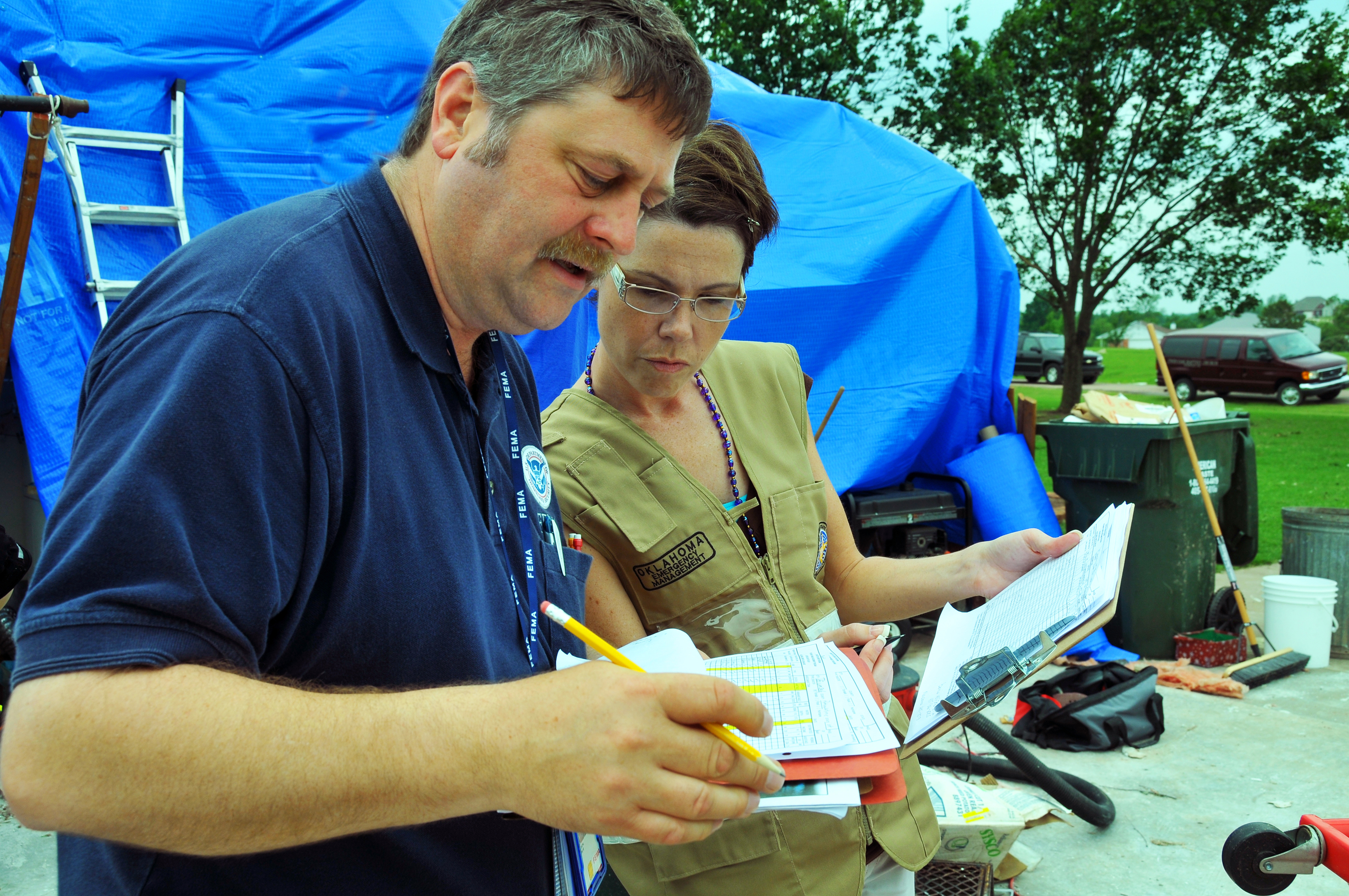 Two inspectors review paperwork outdoors near a blue tarp at a storm-damaged property.