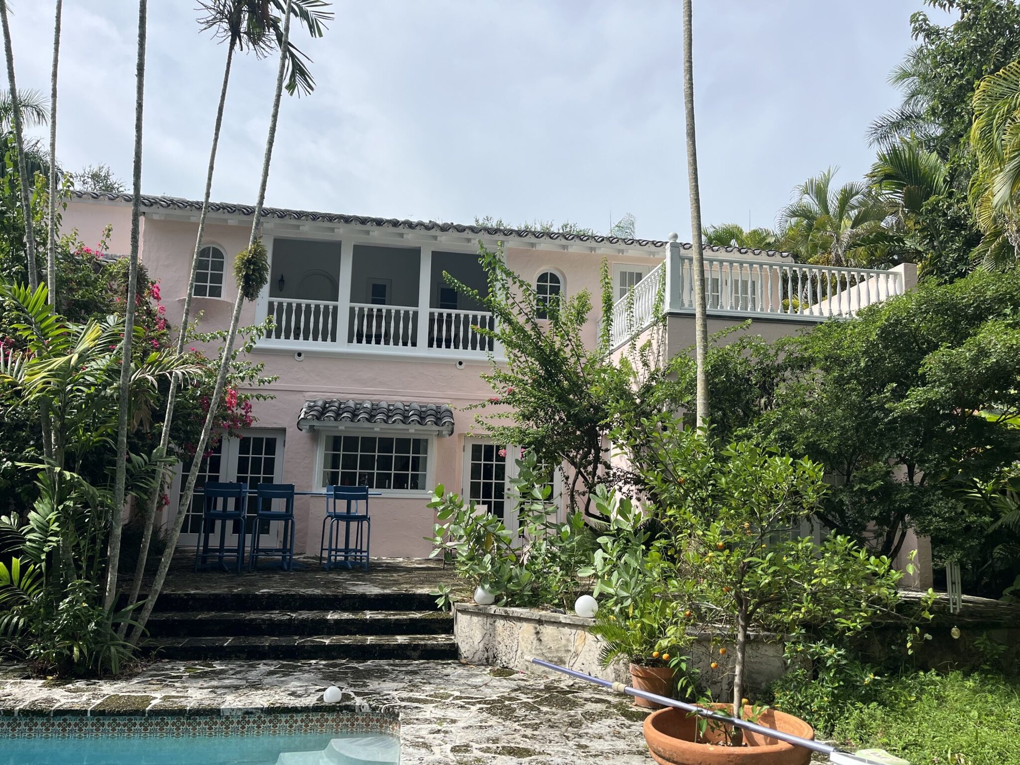 Two-story coral-pink stucco house with white balconies, arched windows, and a pool surrounded by palm trees.