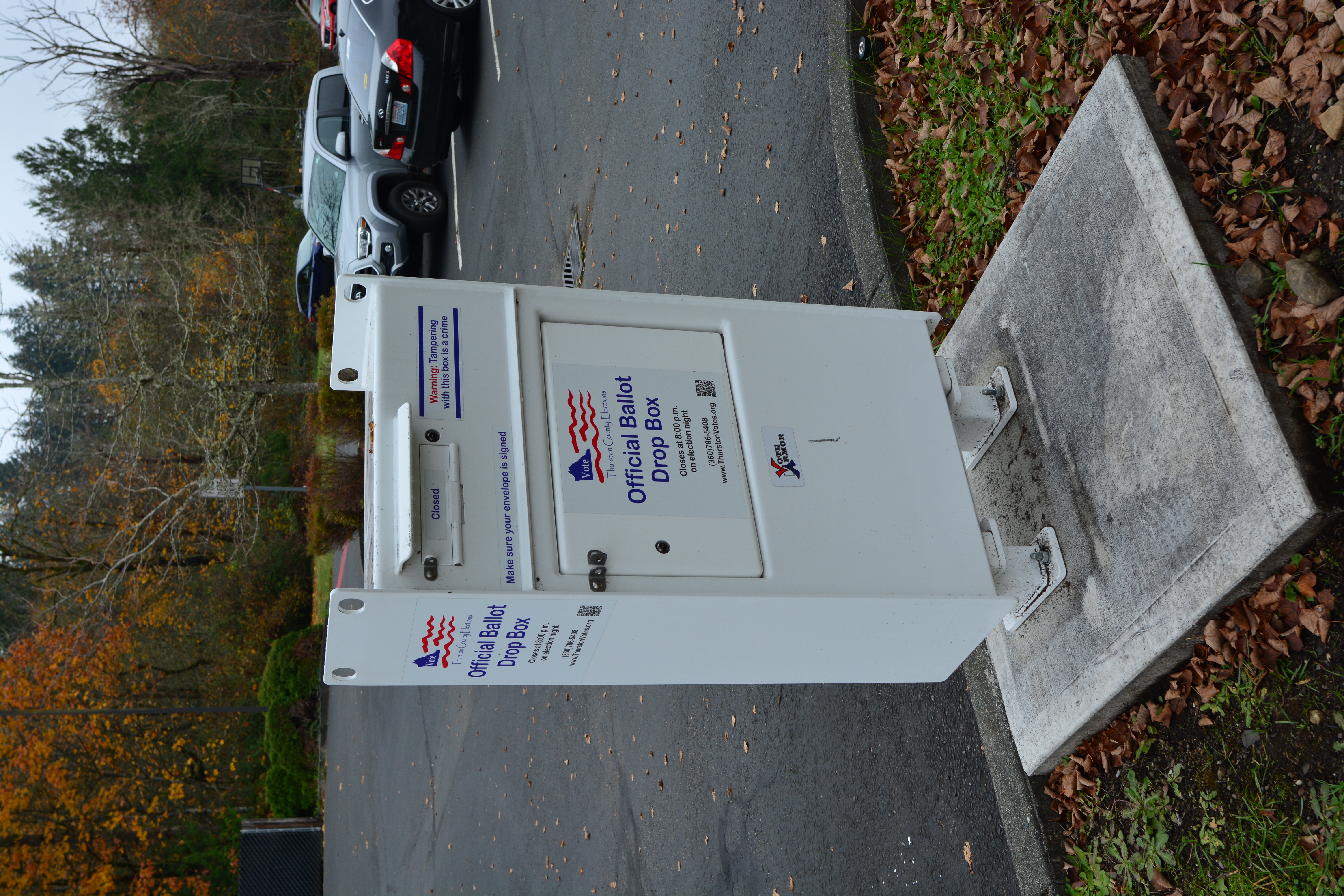 White official ballot drop box on a concrete pad in a parking lot, surrounded by autumn foliage.