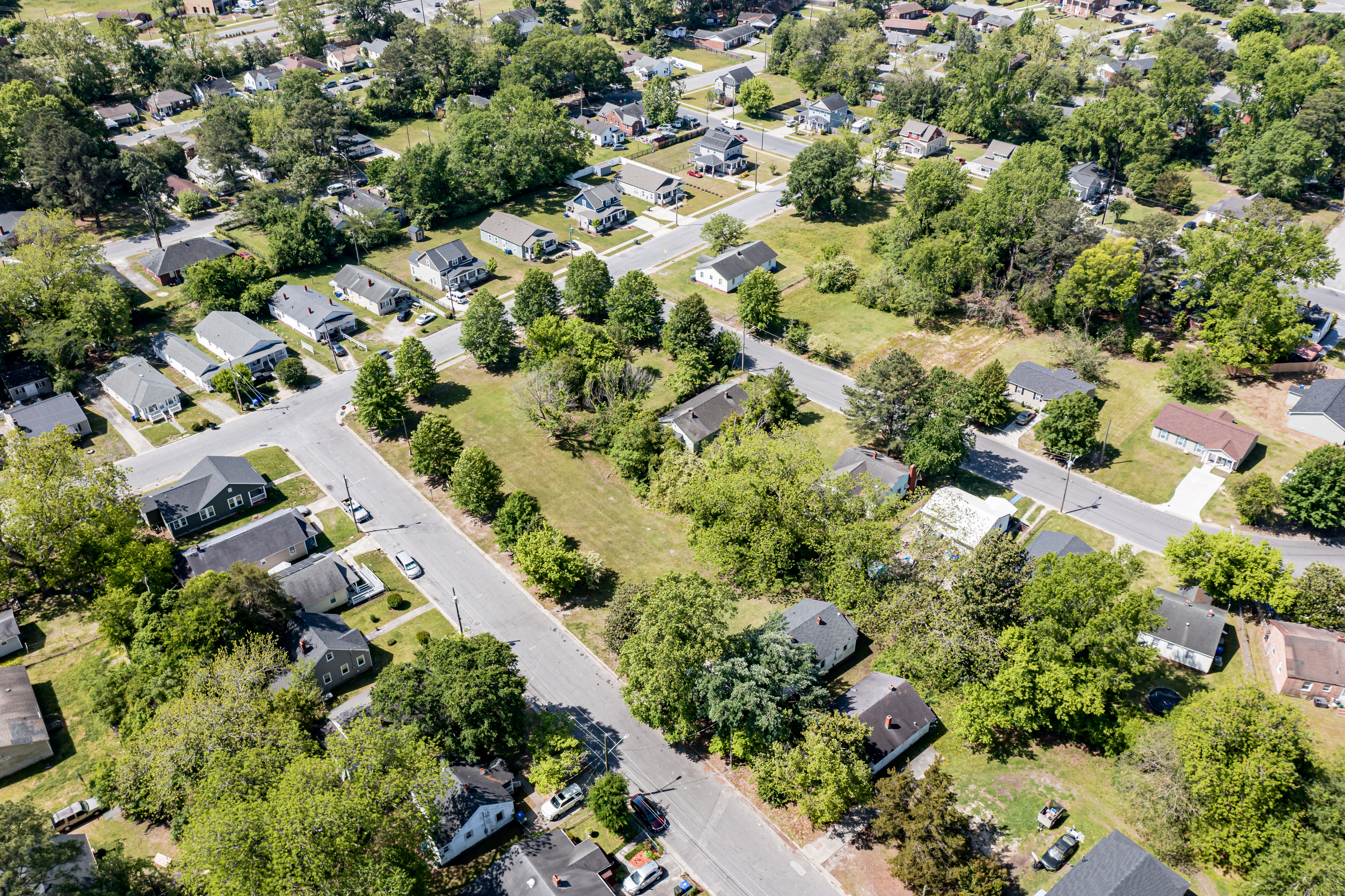 Aerial view of a suburban neighborhood with tree-lined streets, homes, and an open grassy lot.