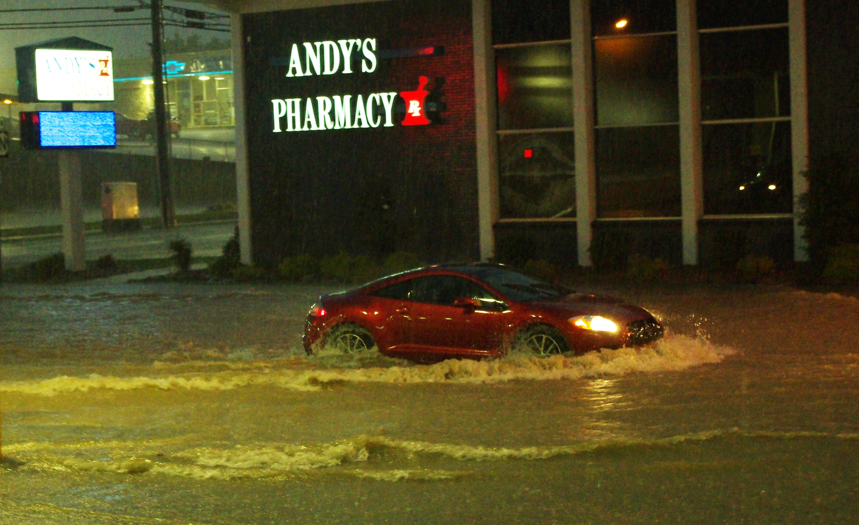 A red car drives through deep floodwaters on a street at night.