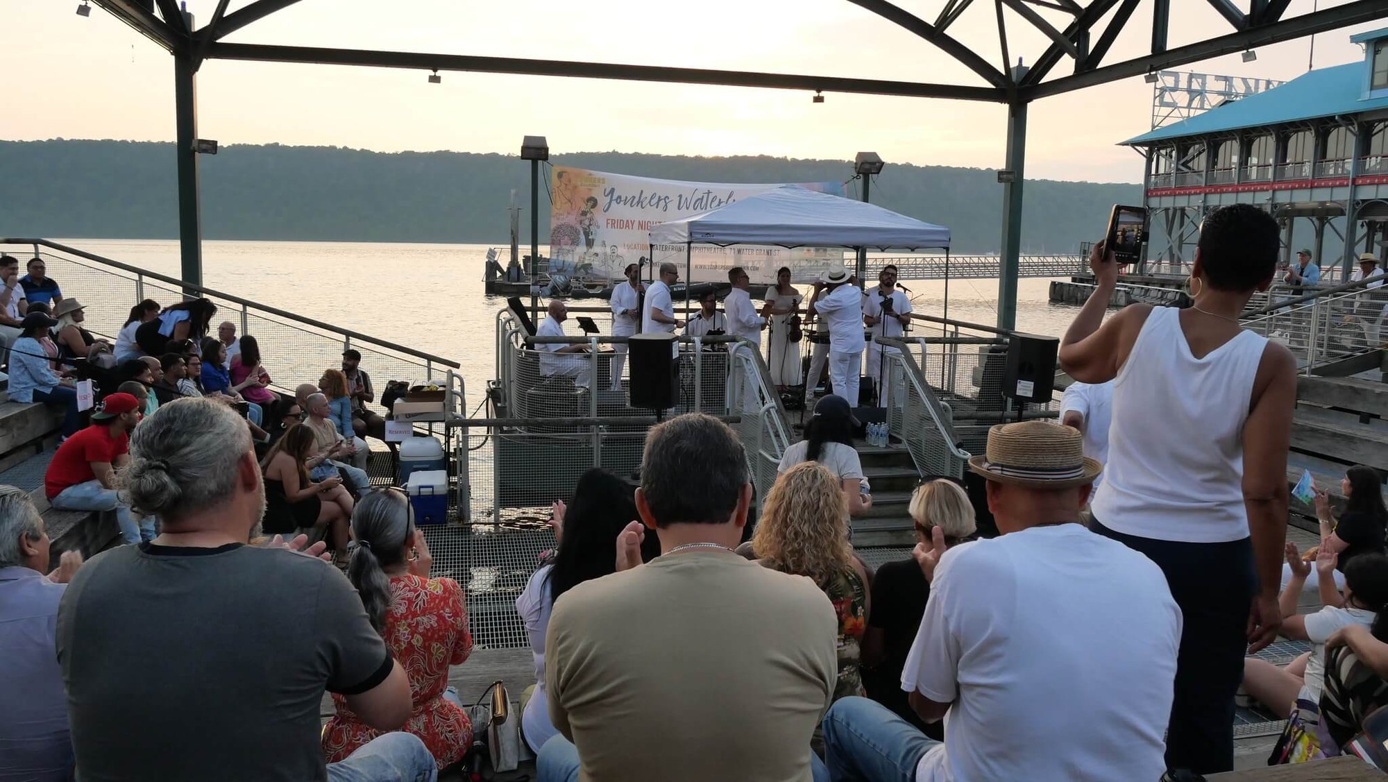 Crowd seated under an open-air pavilion watching a live band perform at dusk near a river.