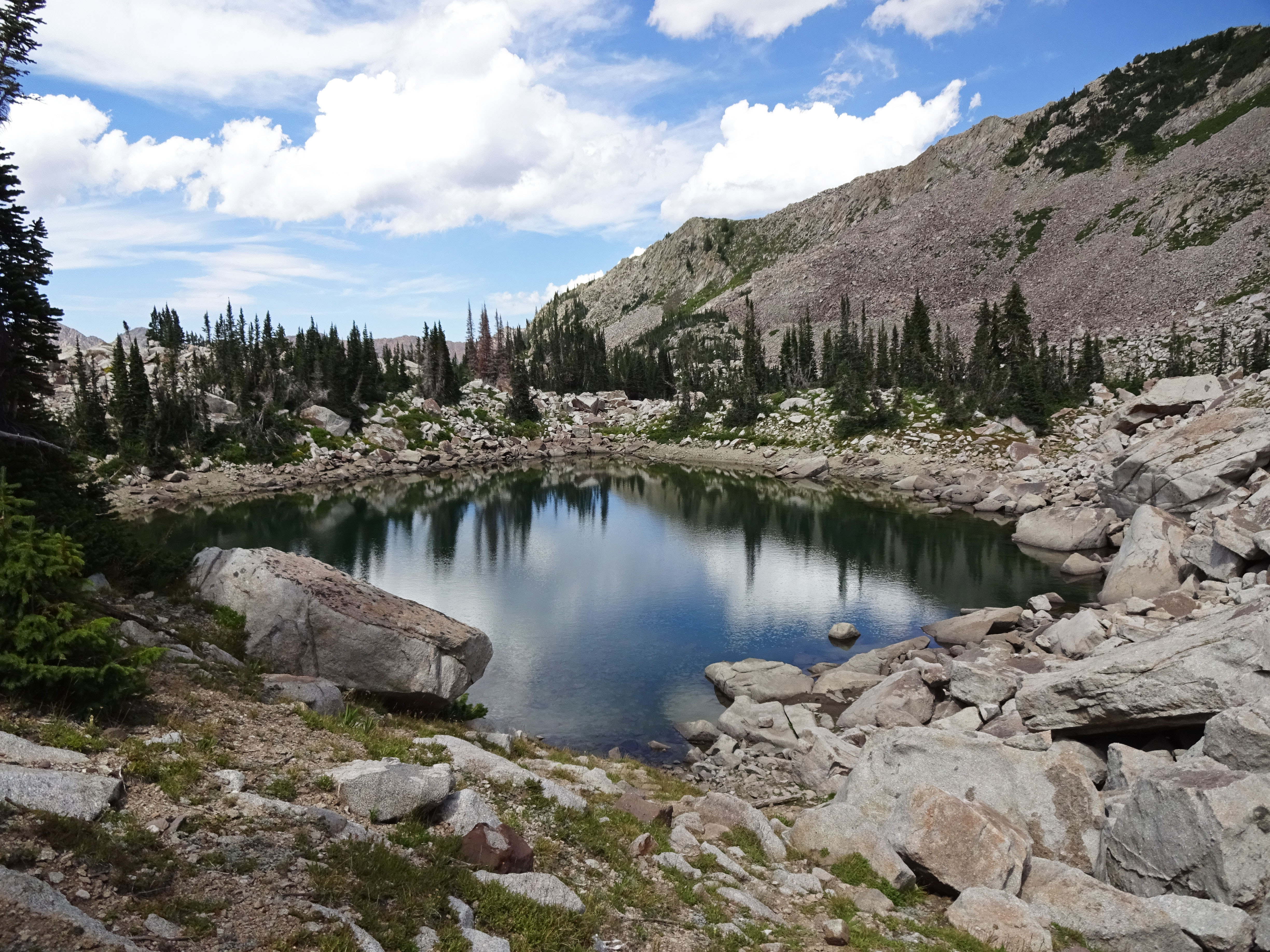 Alpine lake surrounded by boulders and conifers beneath cumulus clouds and rocky peaks.