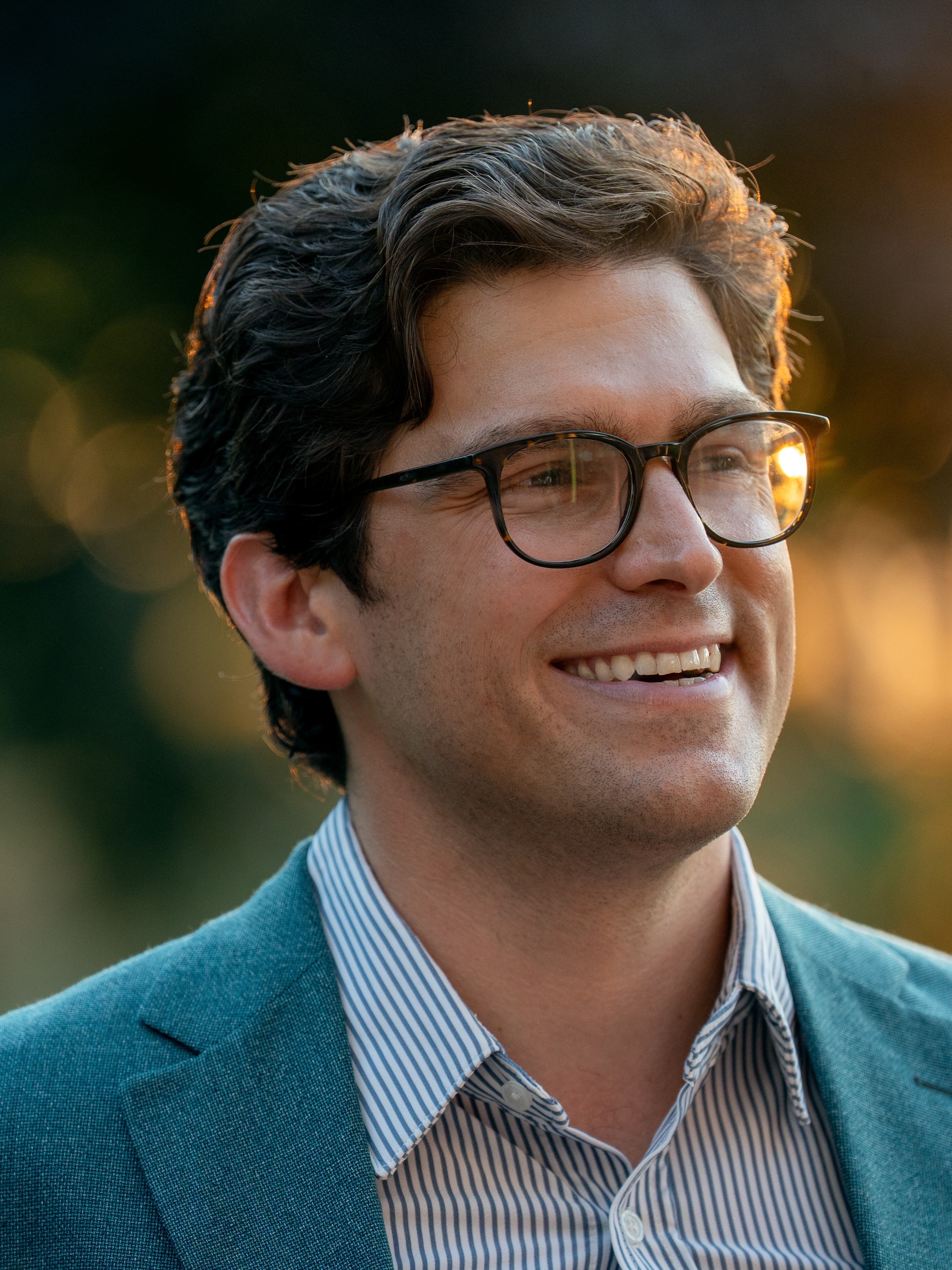 Man with dark hair and glasses wearing a teal blazer, smiling outdoors.