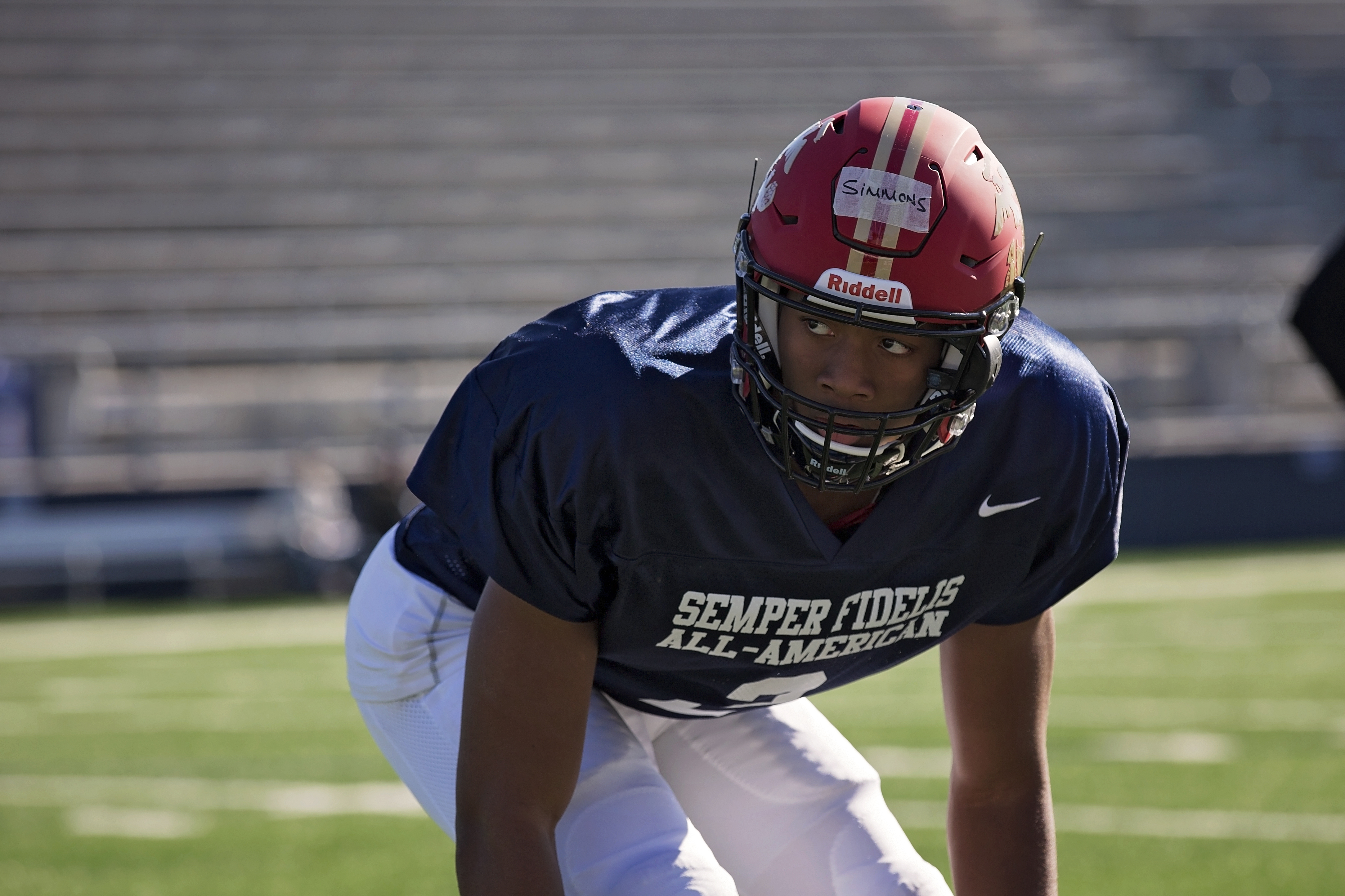 A football player in a red helmet crouches in a pre-snap stance on a practice field.