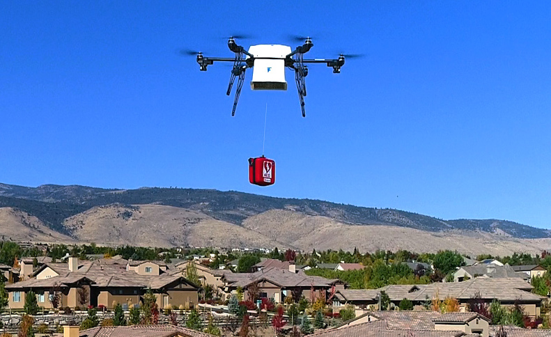 A white quadcopter drone carries a red package suspended by a tether over a suburban neighborhood.