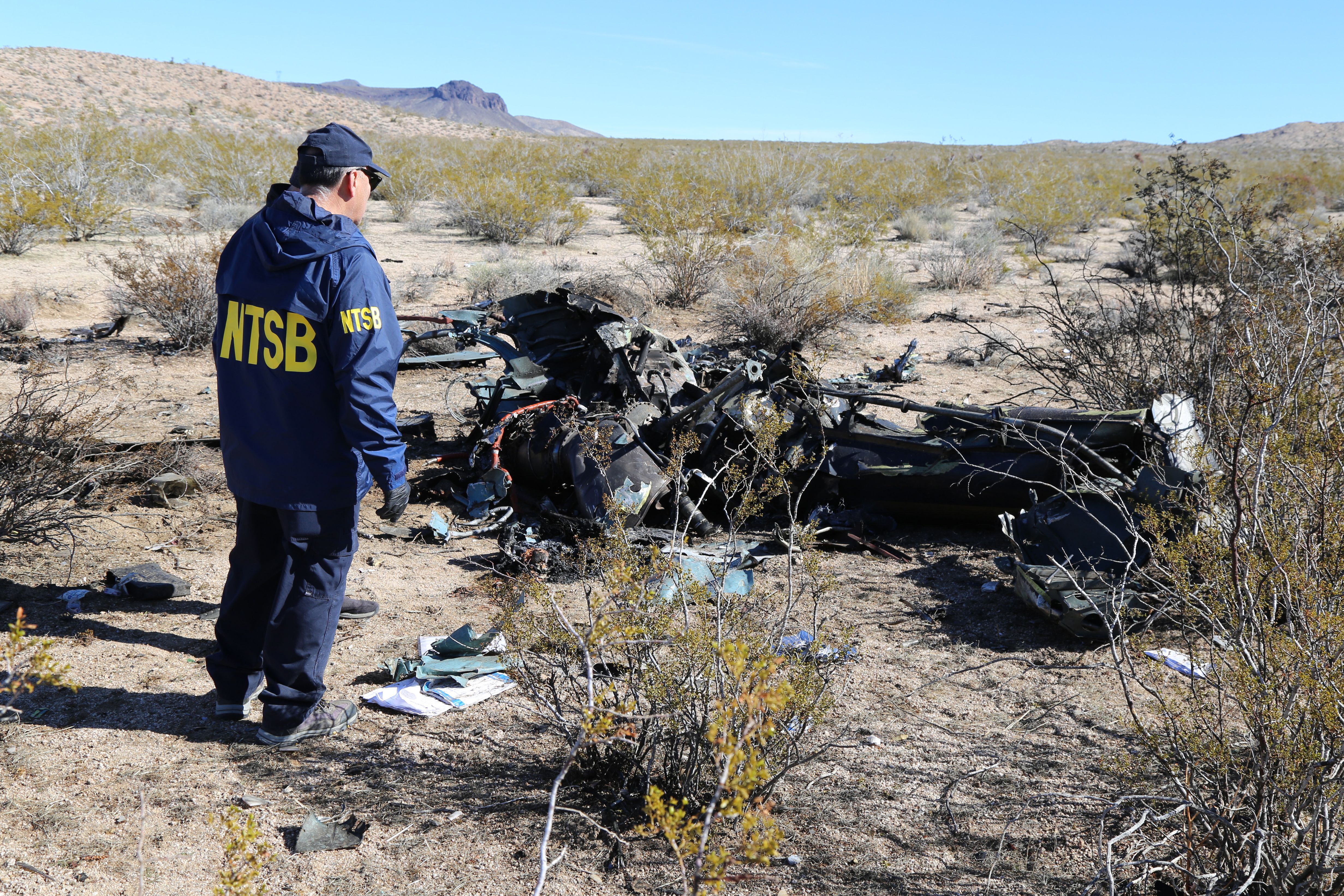 NTSB investigator in blue jacket examining burned helicopter wreckage in desert scrubland.