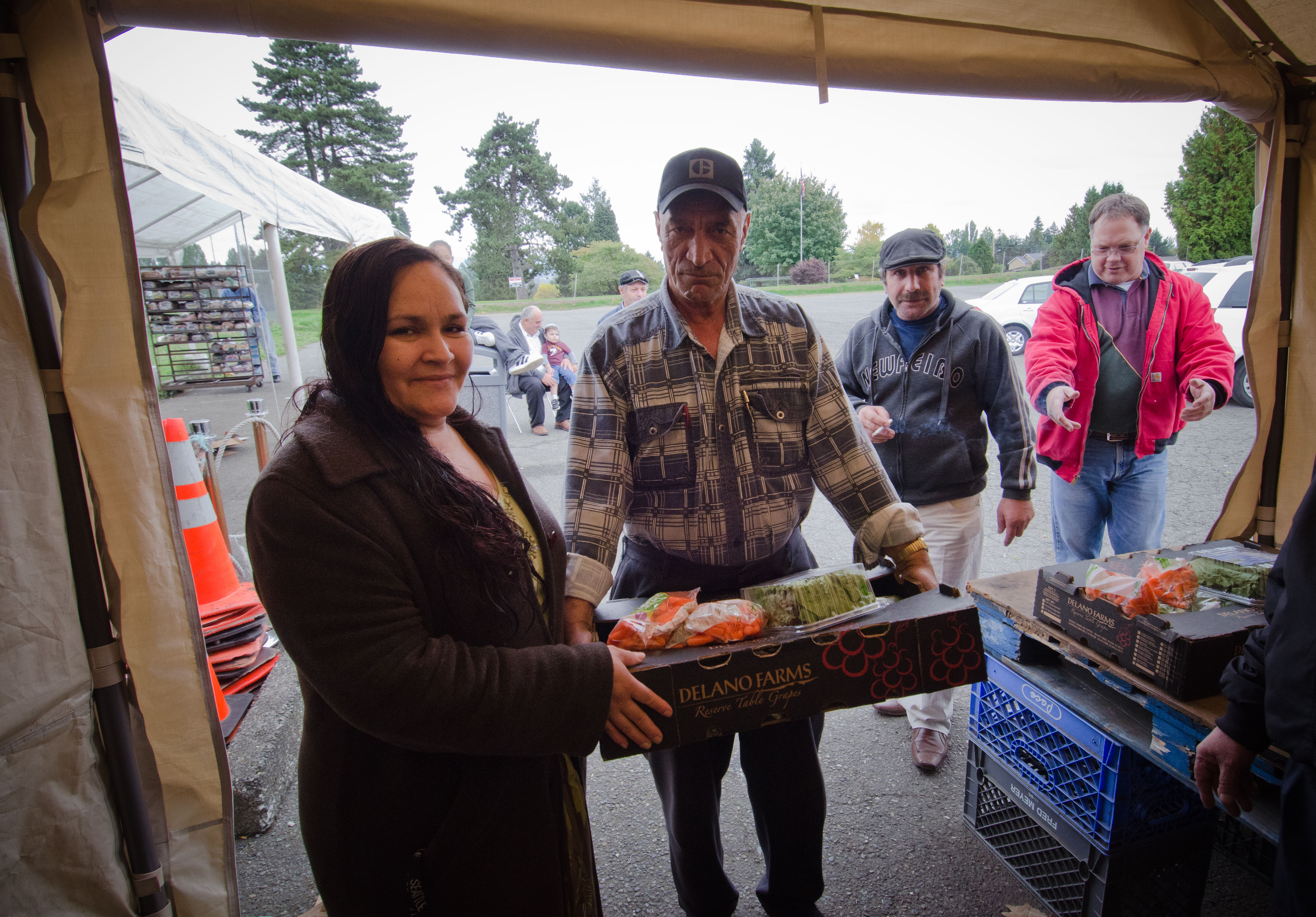 A volunteer hands a box of produce to a recipient at an outdoor distribution tent.