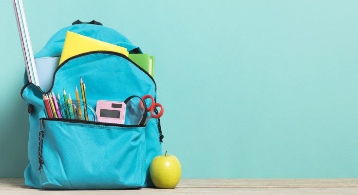 A blue backpack stuffed with pencils, scissors, rulers, and folders next to a green apple on a wooden surface.