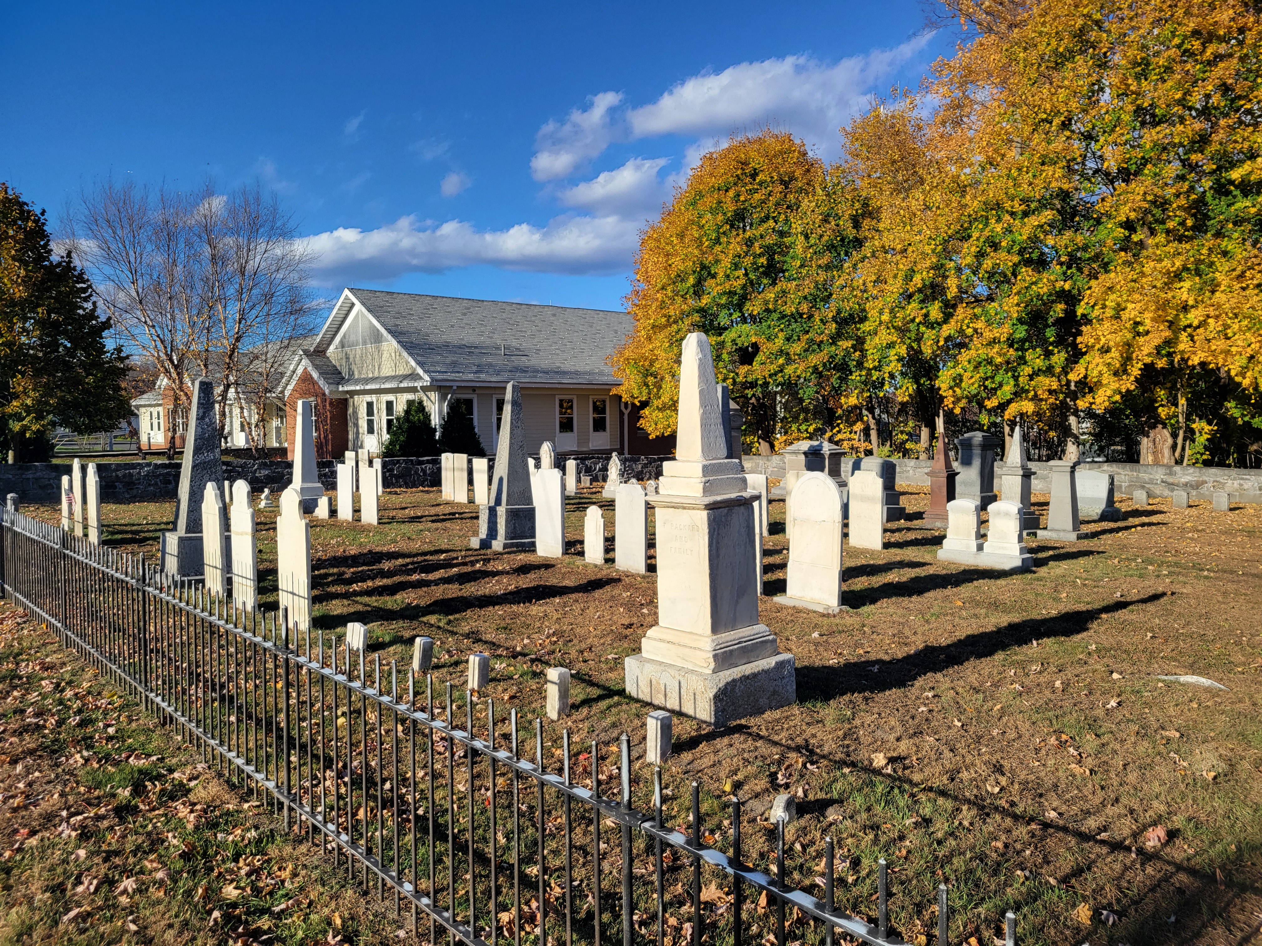 Old gravestones and an obelisk behind a black iron fence with autumn trees.