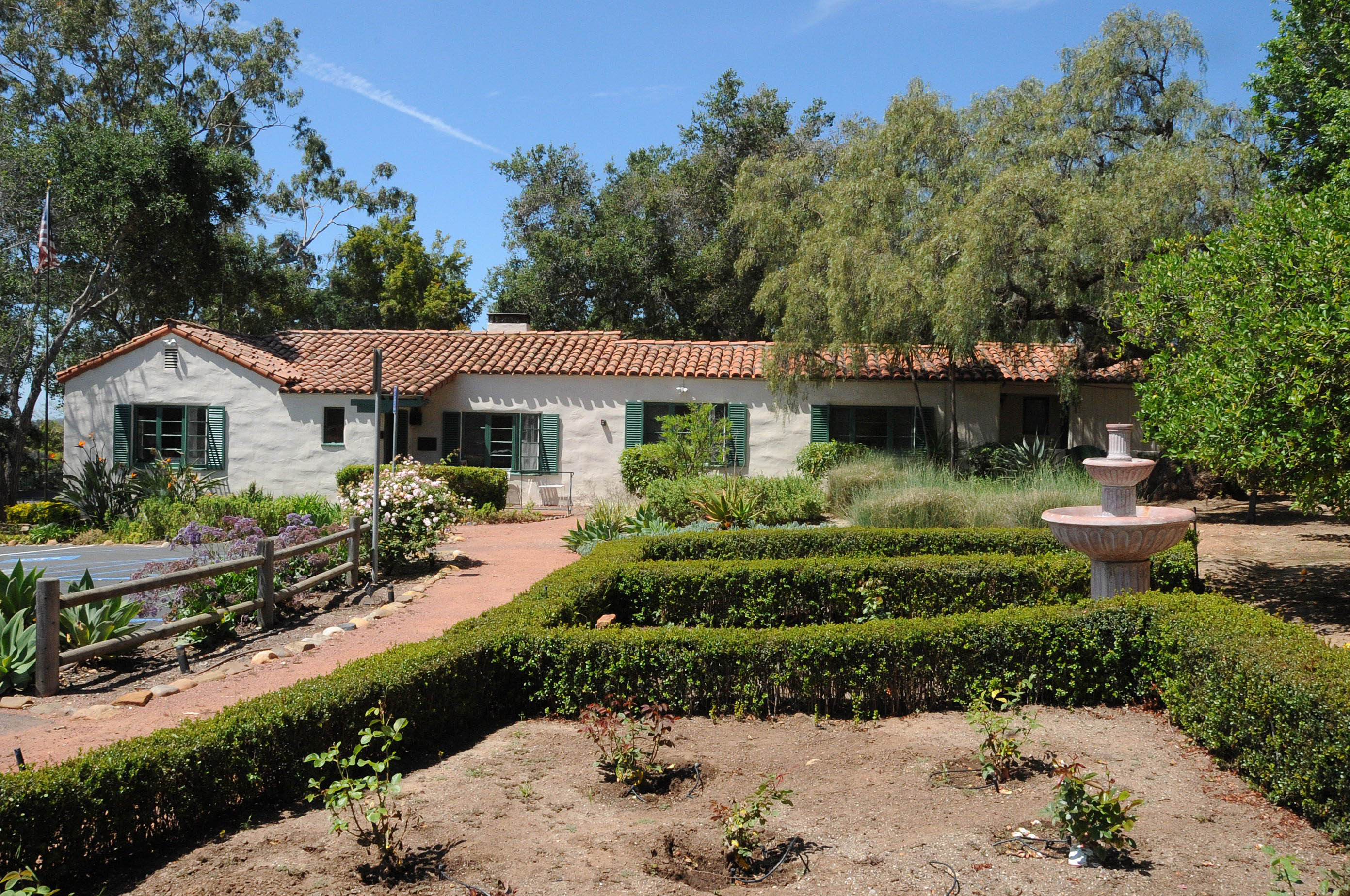 Single-story Spanish Colonial Revival building with red tile roof and formal hedged gardens.