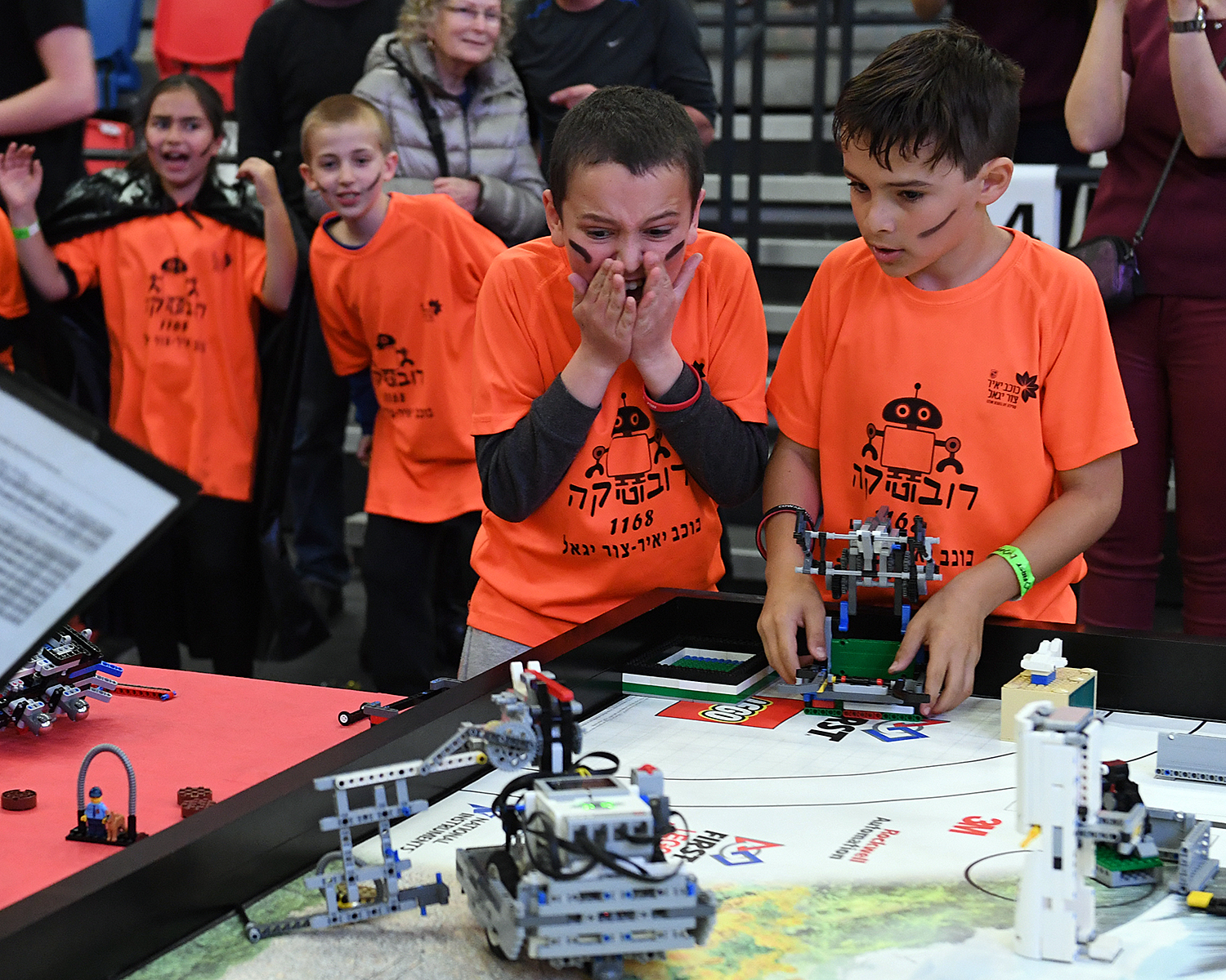 Children in matching orange shirts react excitedly at a LEGO robotics competition table.