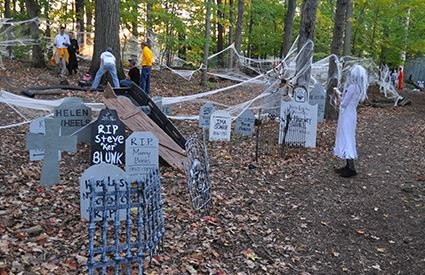 Fake tombstones and cobwebs decorate a wooded outdoor Halloween scene with a costumed figure.