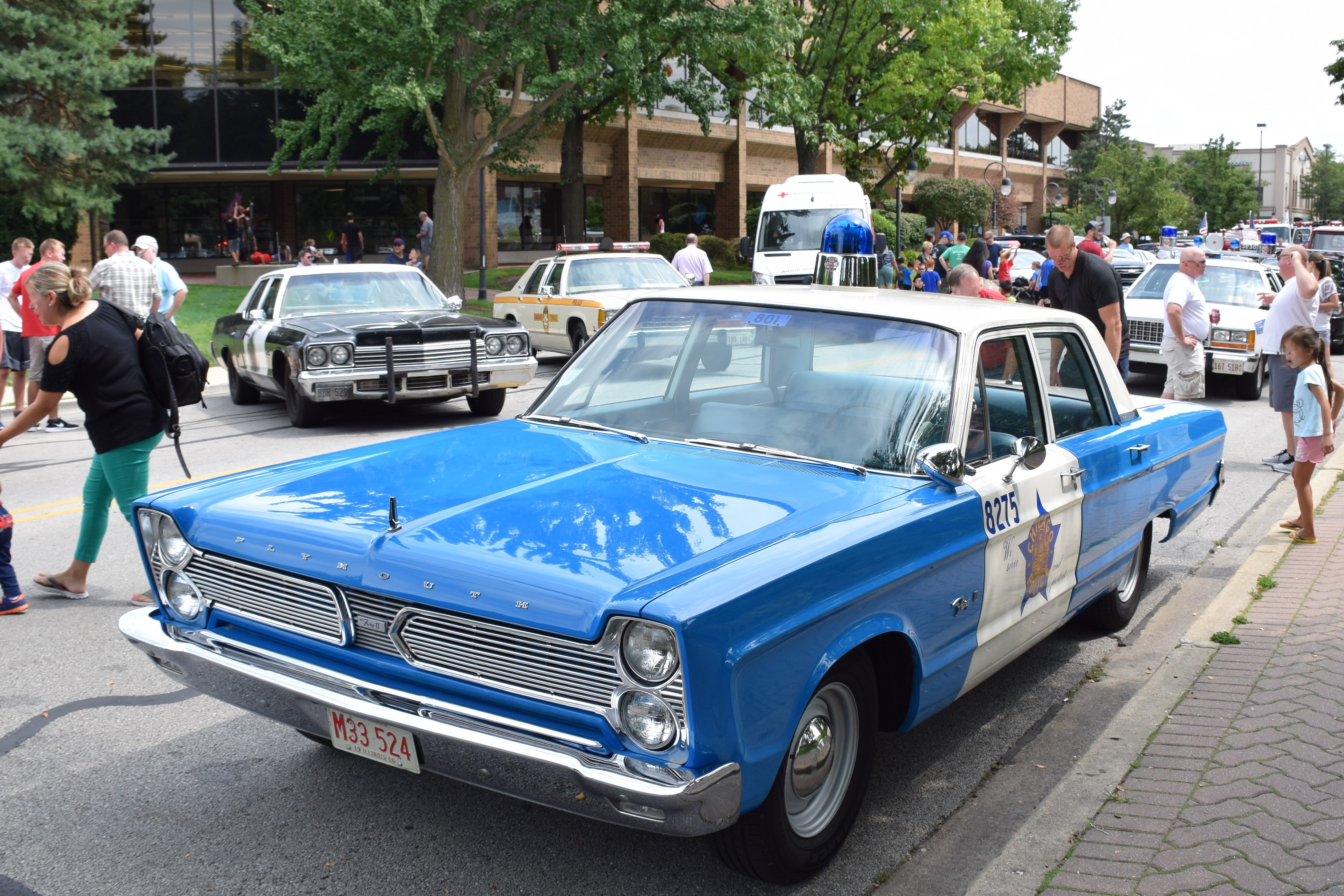 Blue and white vintage police car with a light bar driving at an outdoor exhibition.