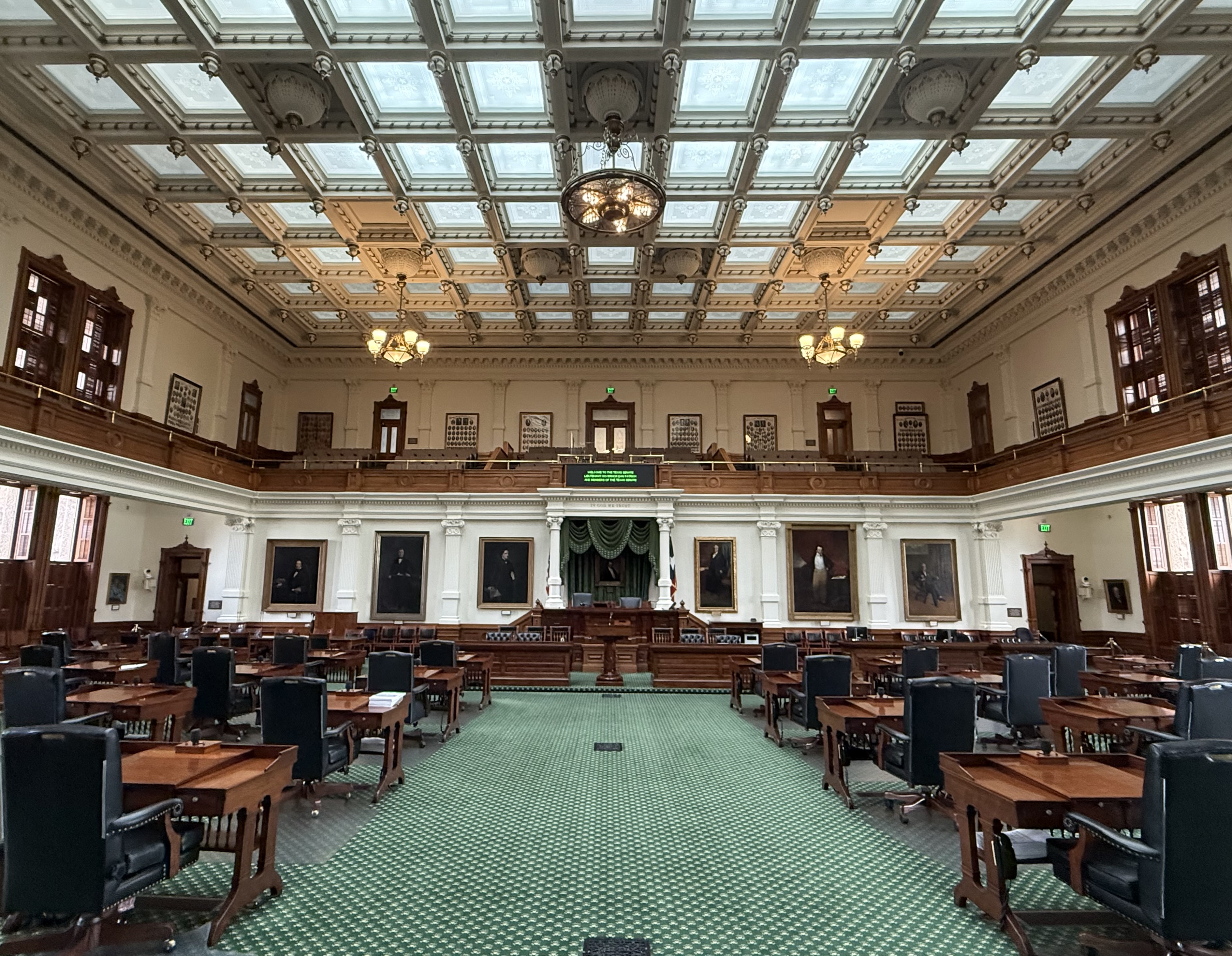 Empty Texas Senate chamber with rows of wooden desks and an ornate coffered ceiling.