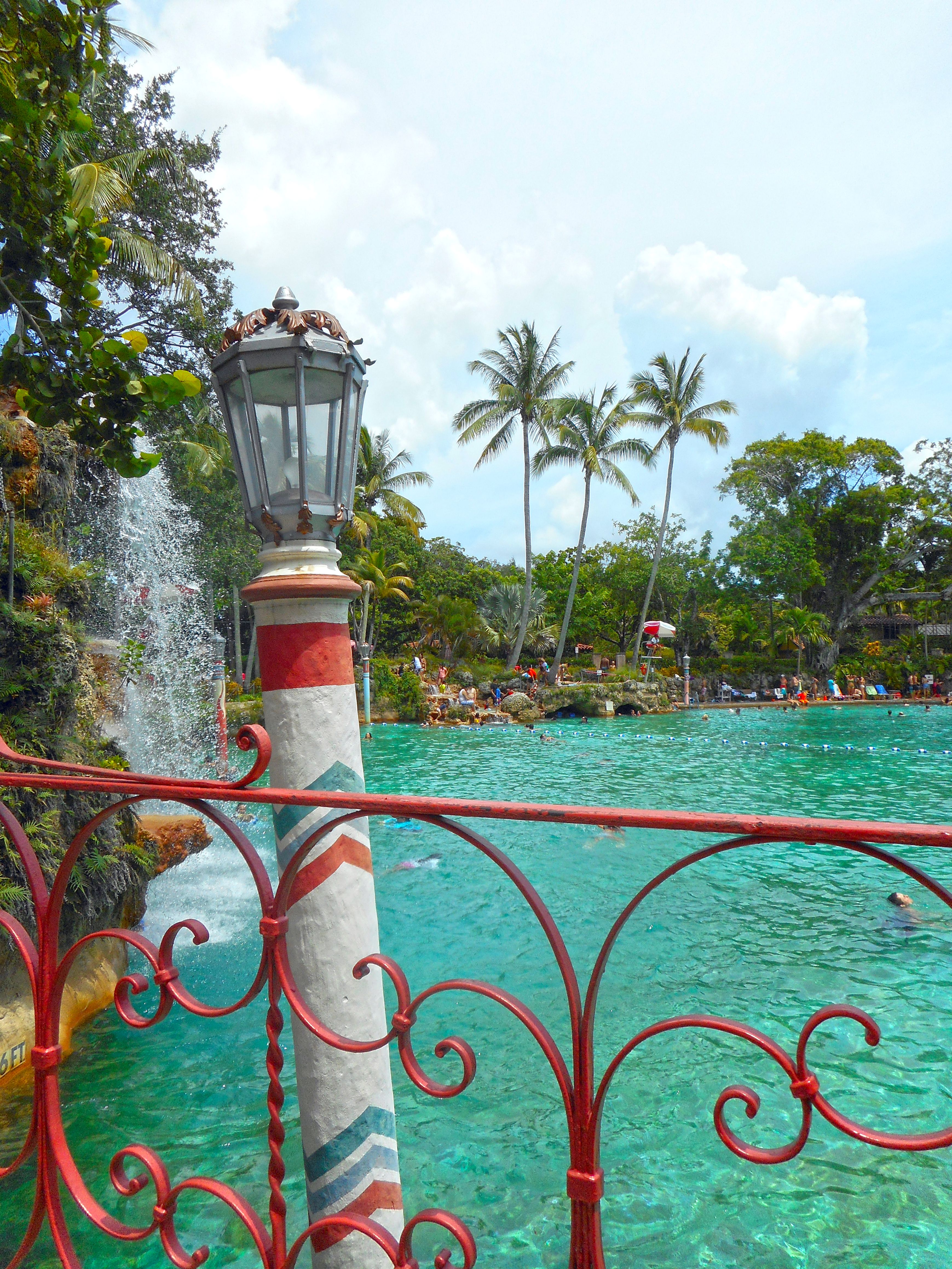 Ornate red iron fence and lamp post framing a turquoise pool with a waterfall and palm trees.
