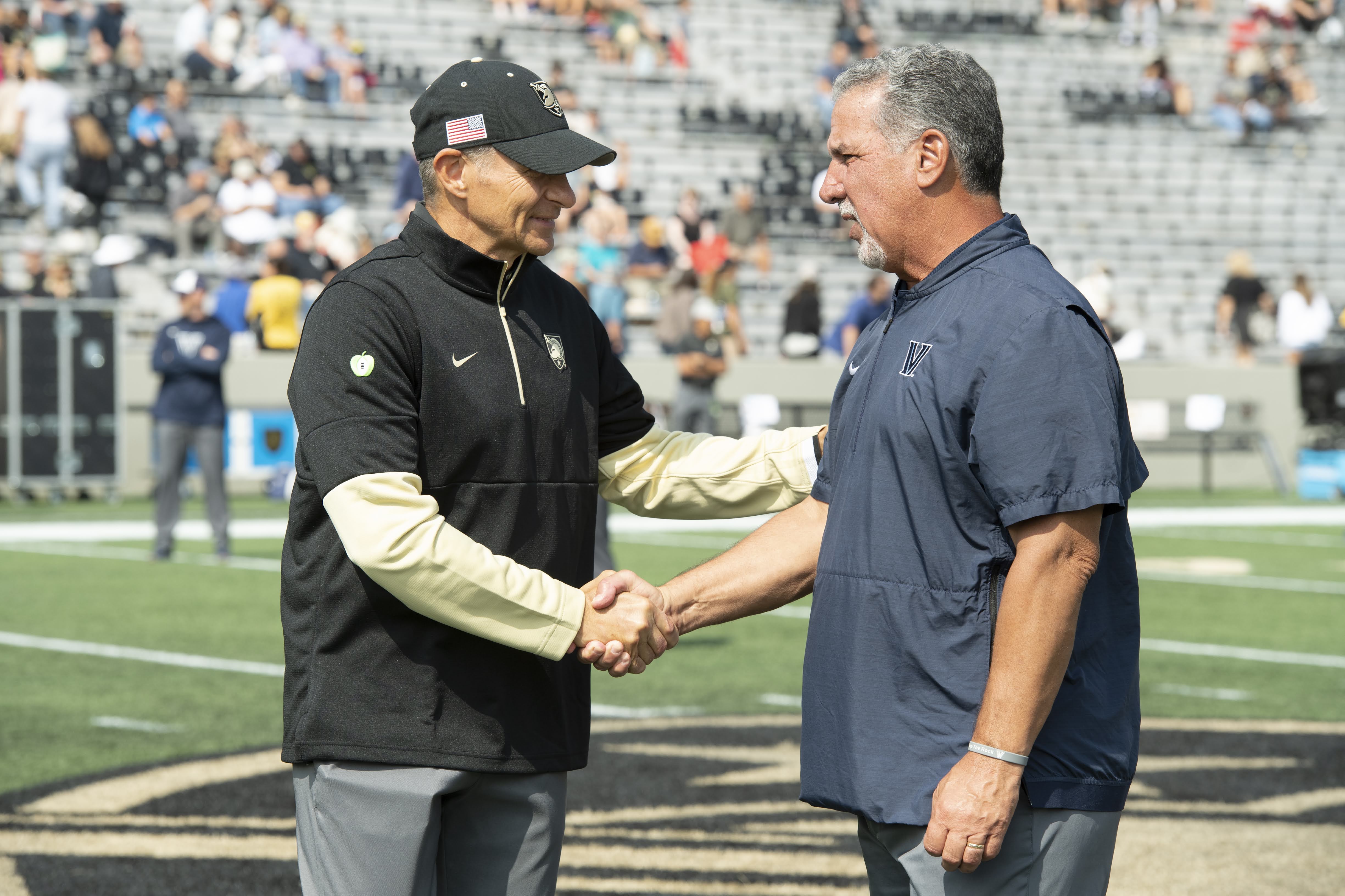 Two football coaches in opposing team attire shake hands on a field before a game.