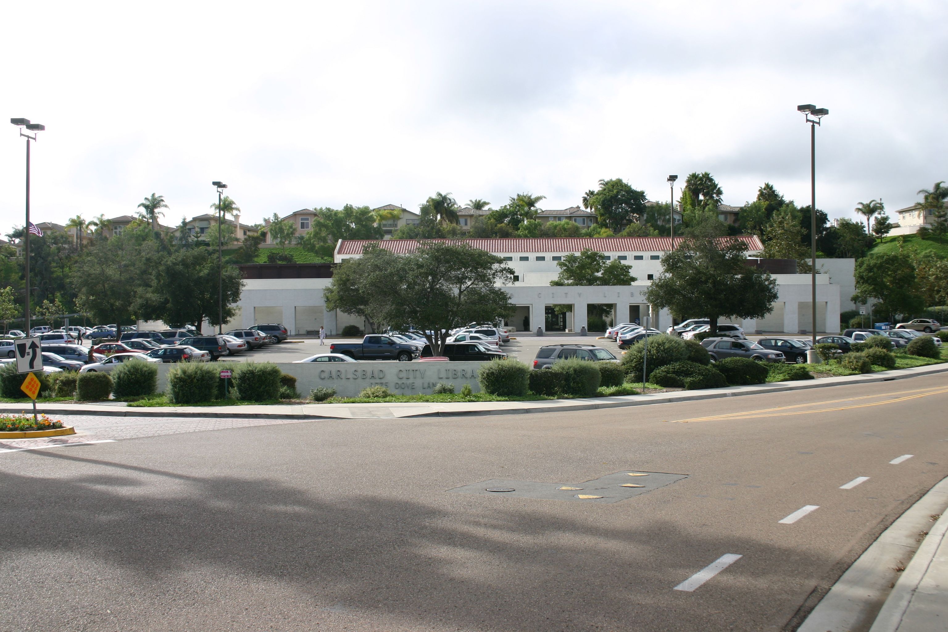 Single-story white building with palm trees and a parking lot full of cars under an overcast sky.