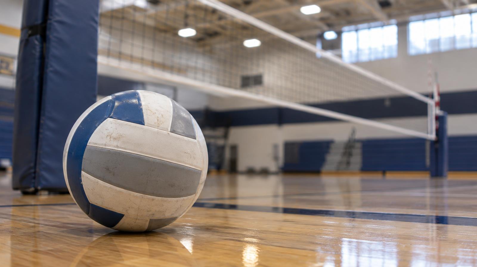 A volleyball sitting on a hardwood floor beside a volleyball net post.