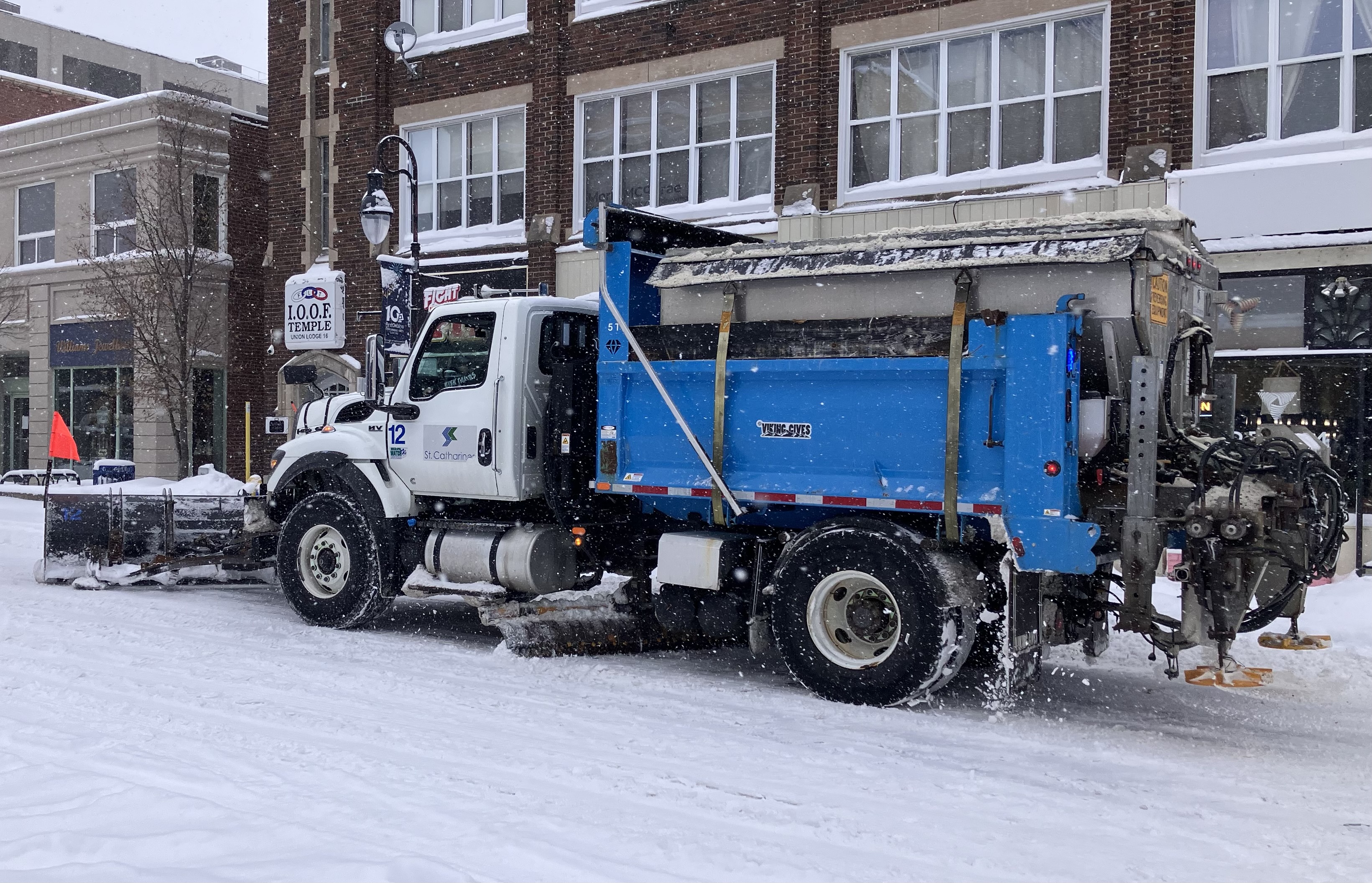 A white and blue municipal snowplow truck with a front plow blade on a snowy street.