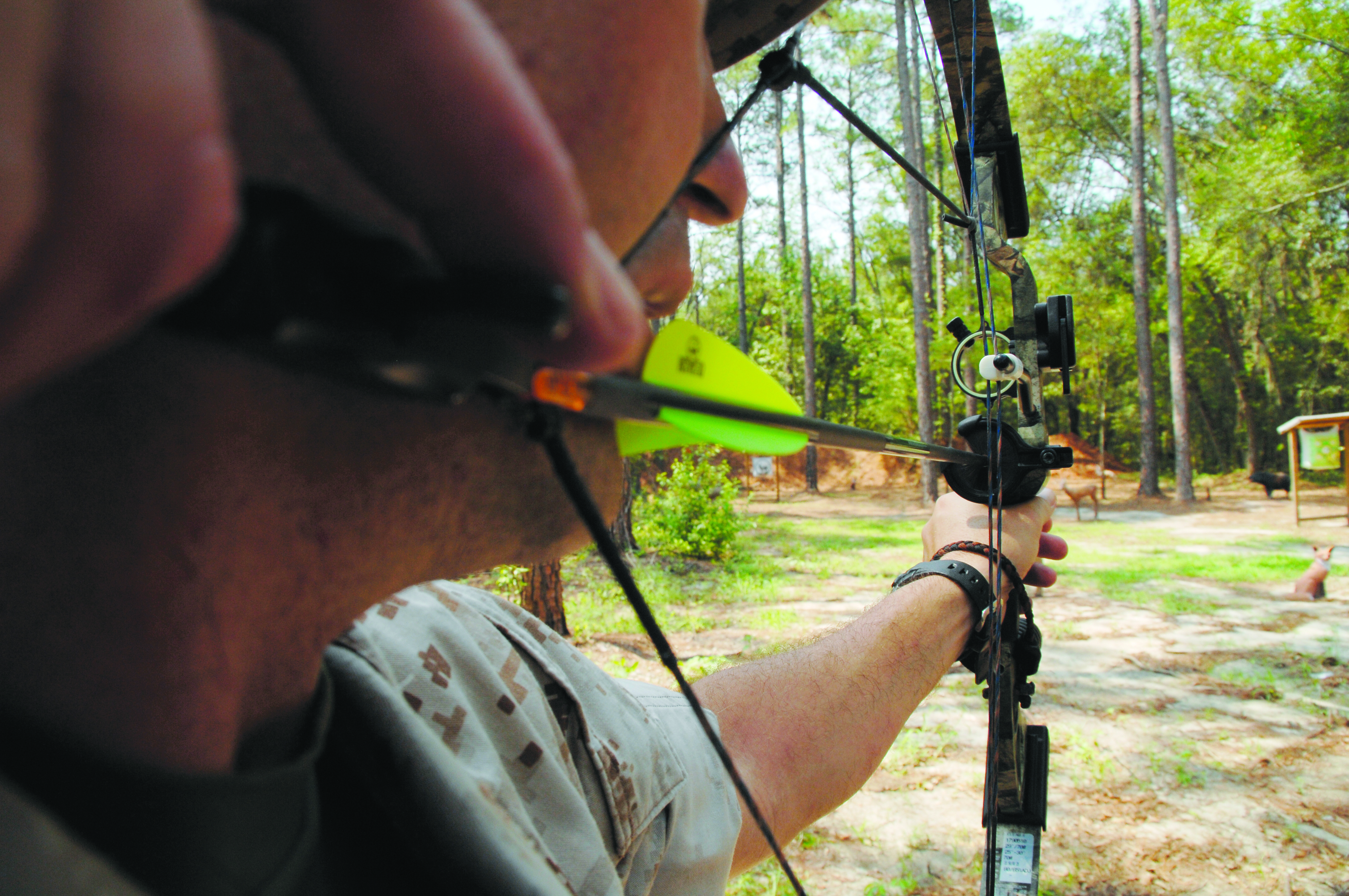 A camouflaged archer draws a compound bow in a wooded outdoor setting.