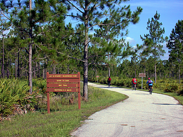 Park entrance sign and paved trail through pine flatwoods with cyclists in the distance.