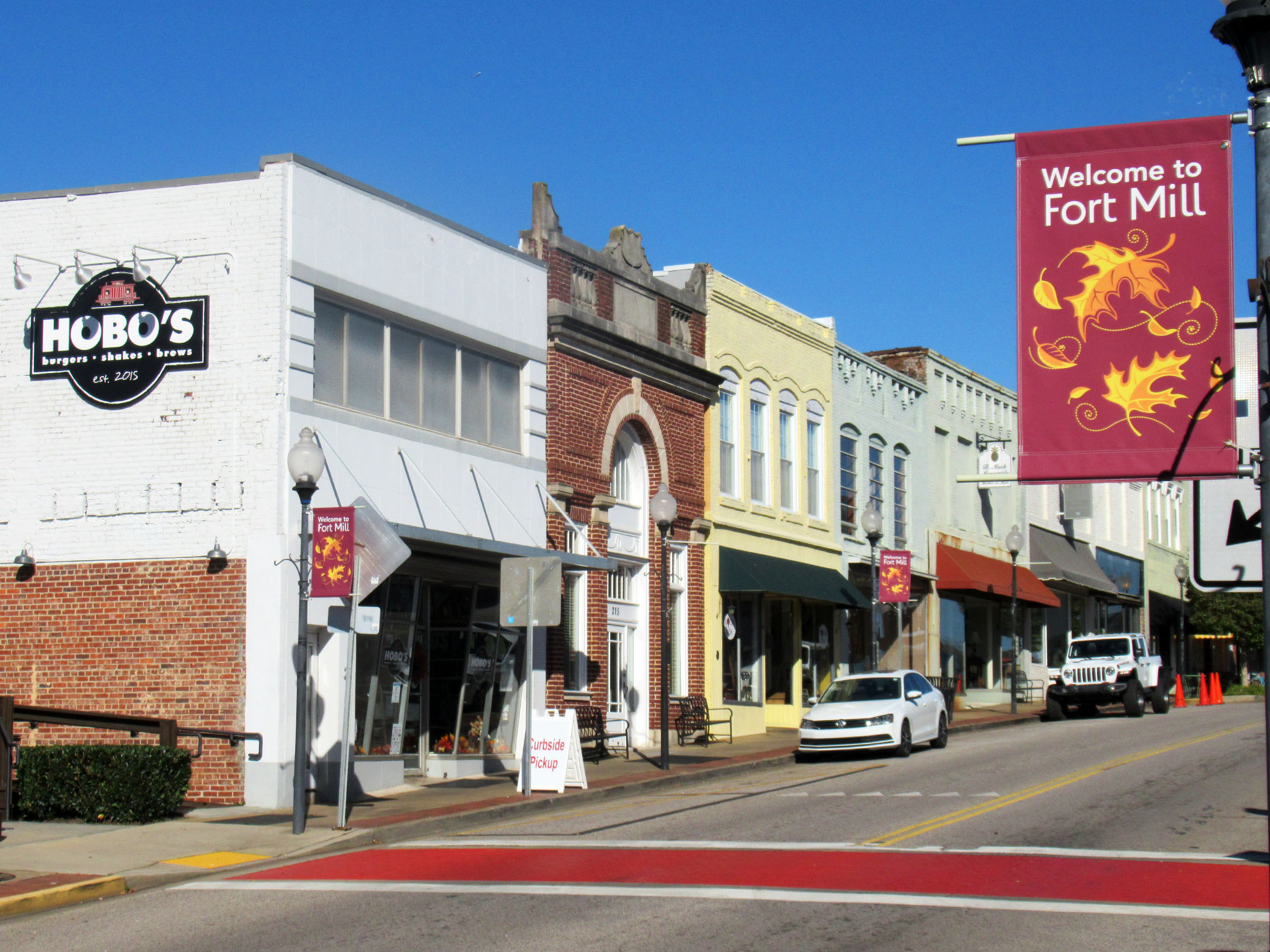 Downtown Fort Mill Main Street with storefronts and a Welcome to Fort Mill banner under a blue sky.
