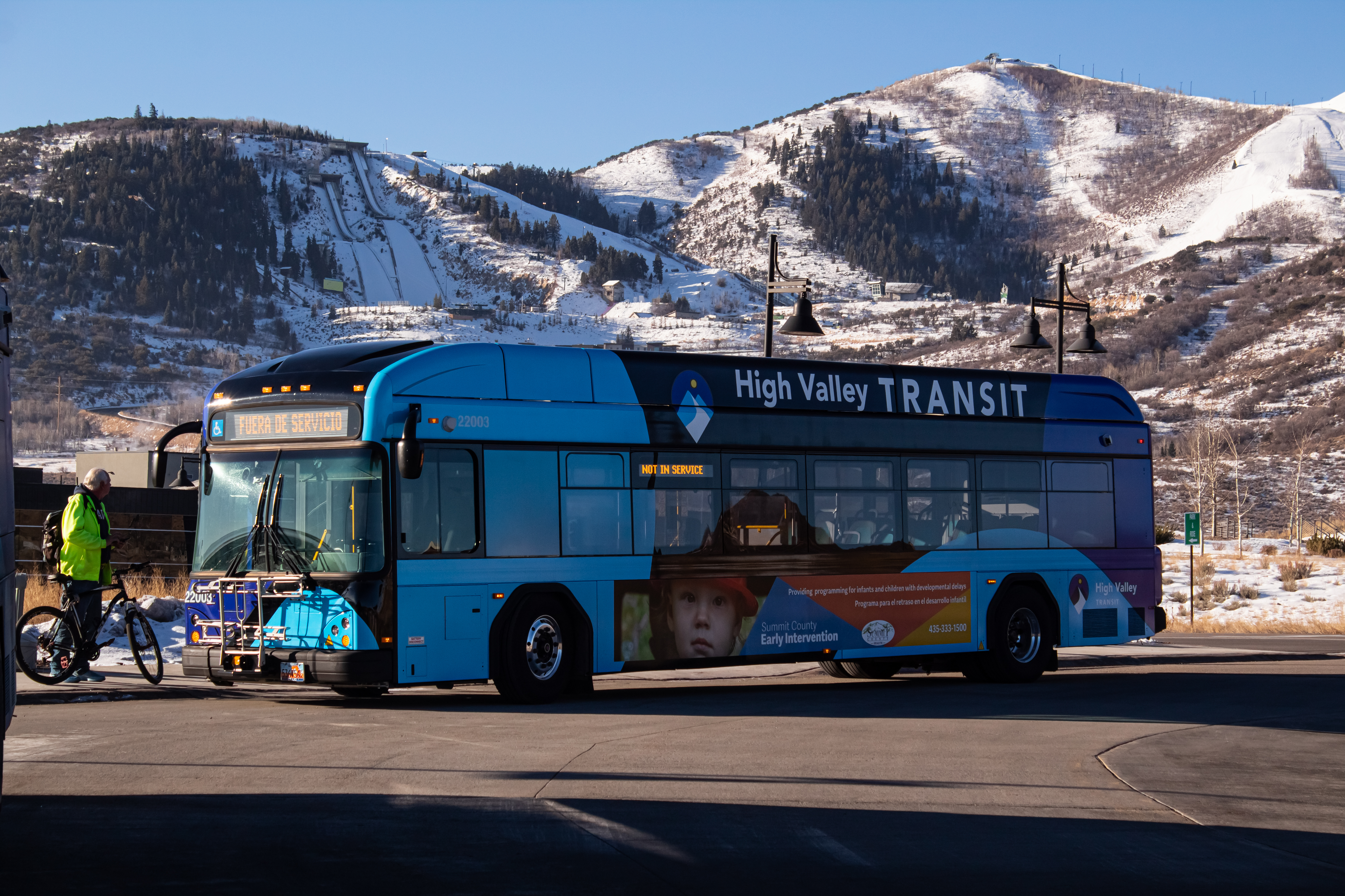 Blue transit bus parked at a transit hub with snow-covered mountains in the background.