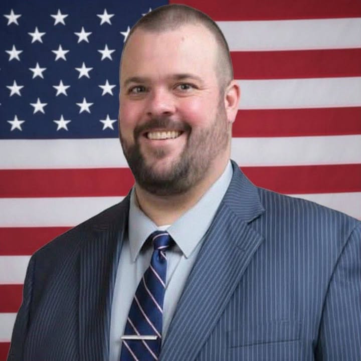 Shanon R. Holm wearing a suit and tie in front of an American flag.