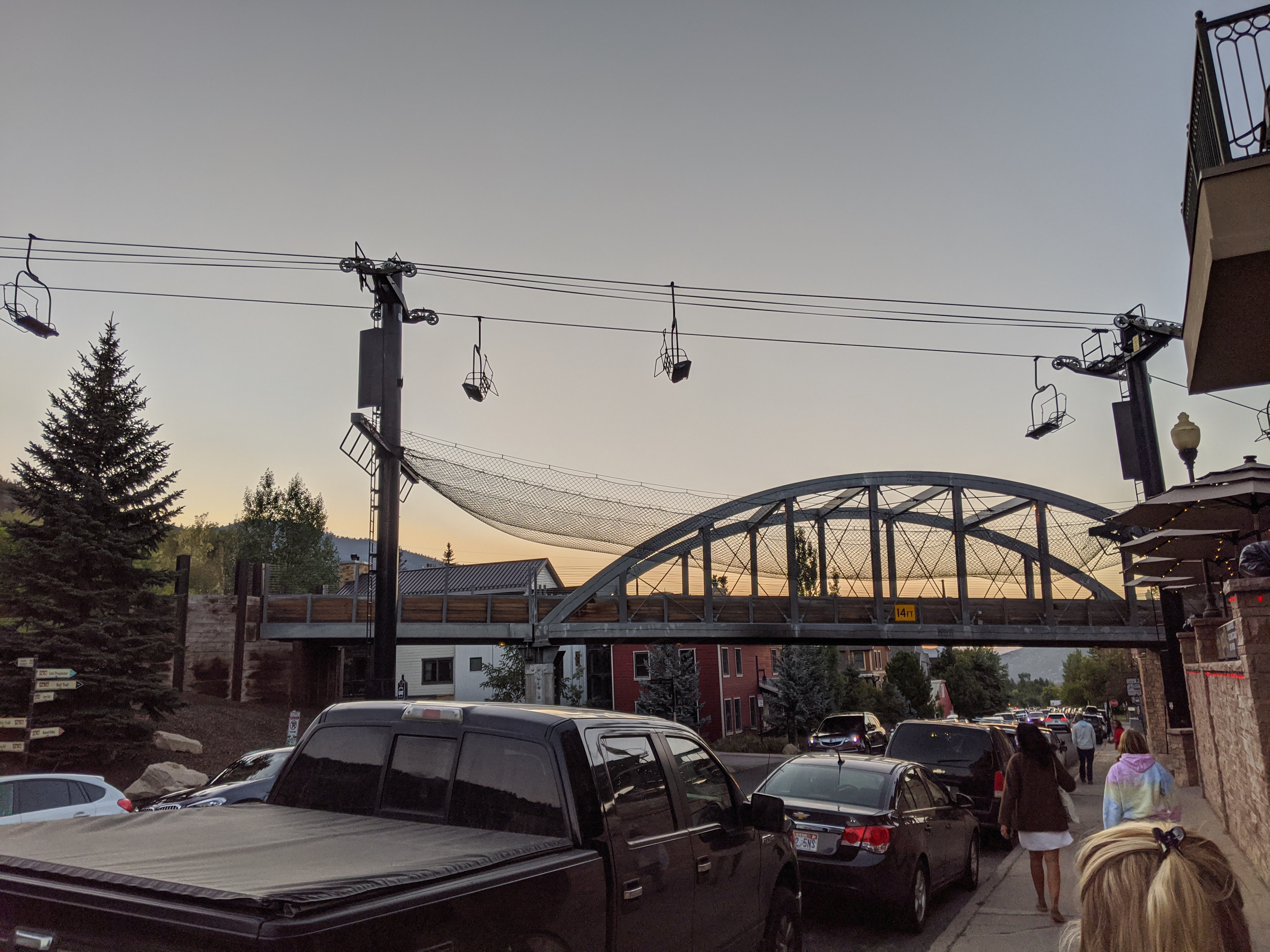 Downtown street at dusk with gondola chairs on overhead cables and an arched bridge.