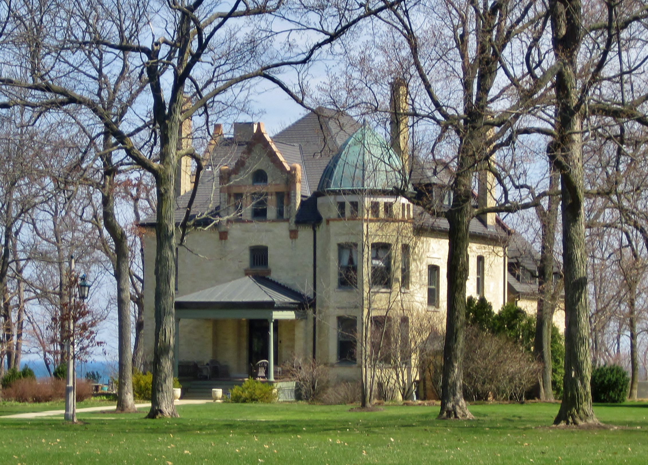 Large Victorian stone mansion with a copper dome roof surrounded by trees on a manicured lawn.