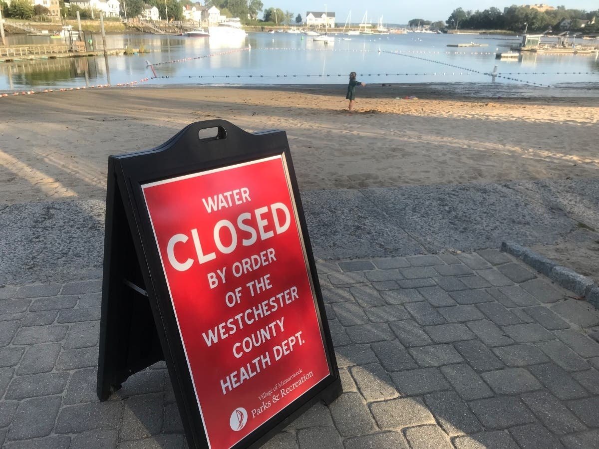 Red A-frame sign reading 'Water Closed by Order of the Westchester County Health Dept.' near a sandy beach and marina.