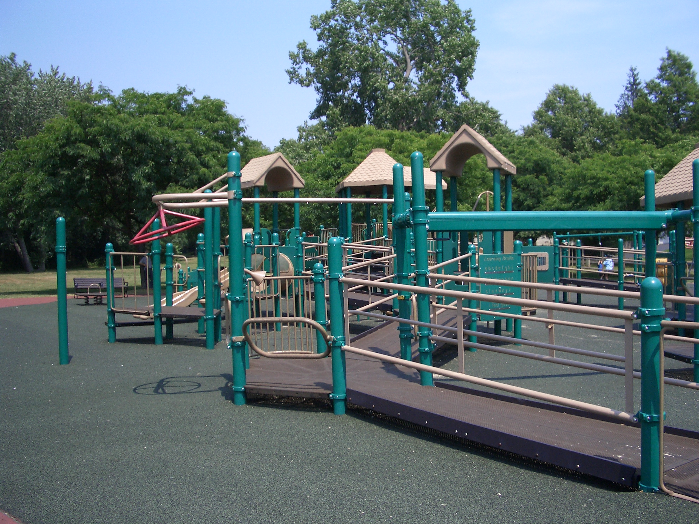 Accessible playground structure with teal ramps, railings, and roofed platforms on rubberized surfacing.