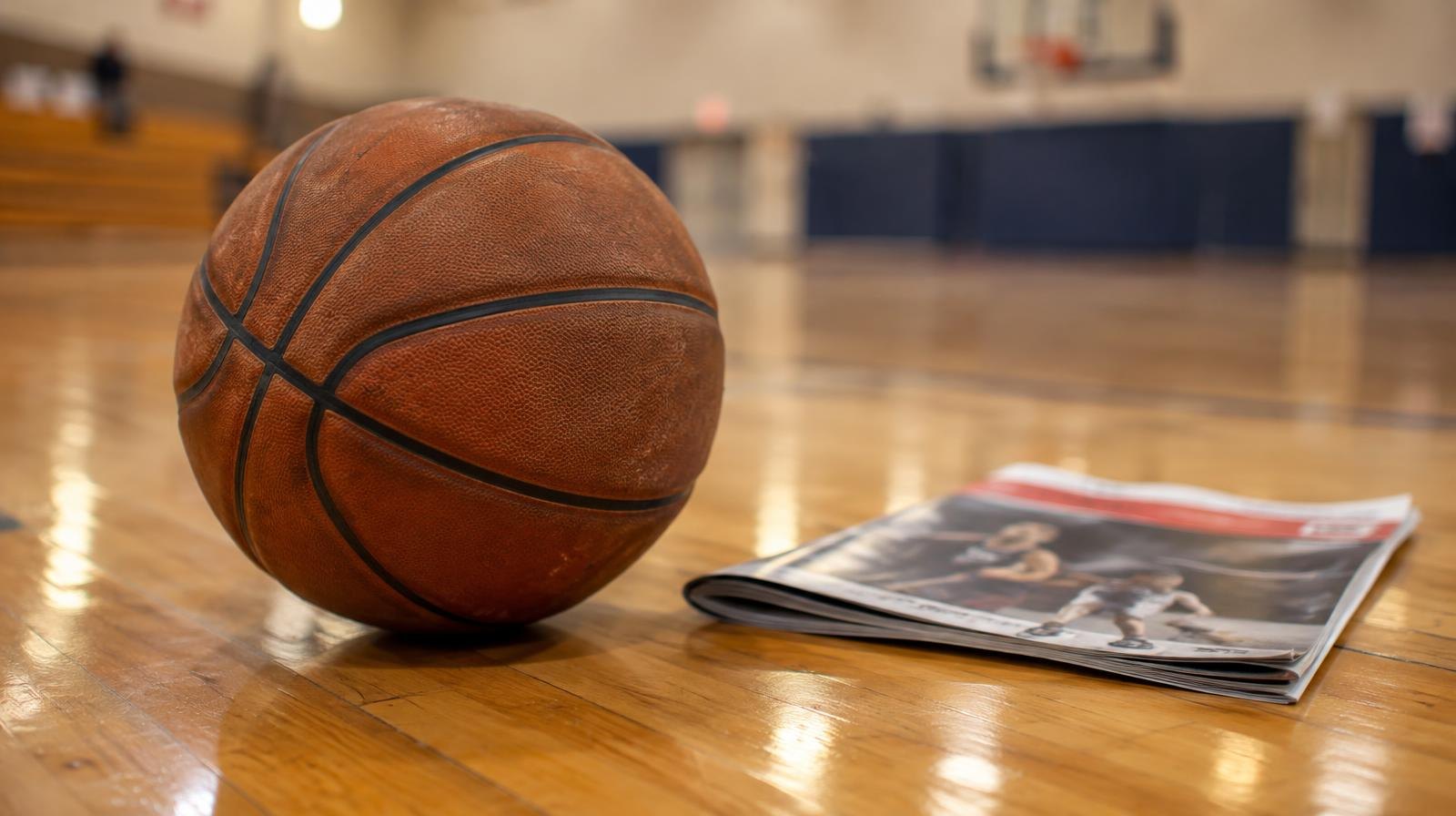 A basketball on a hardwood gym floor next to a folded sports magazine.