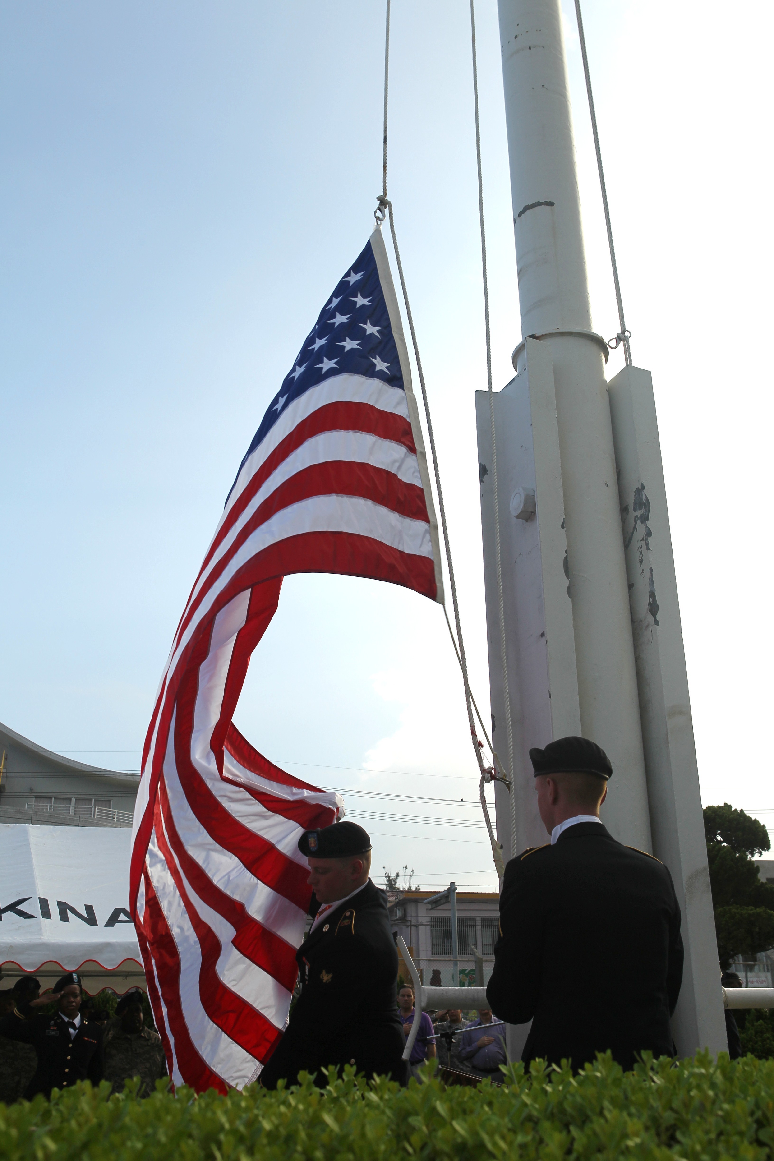 Two uniformed soldiers lower an American flag on a tall flagpole at a ceremony.