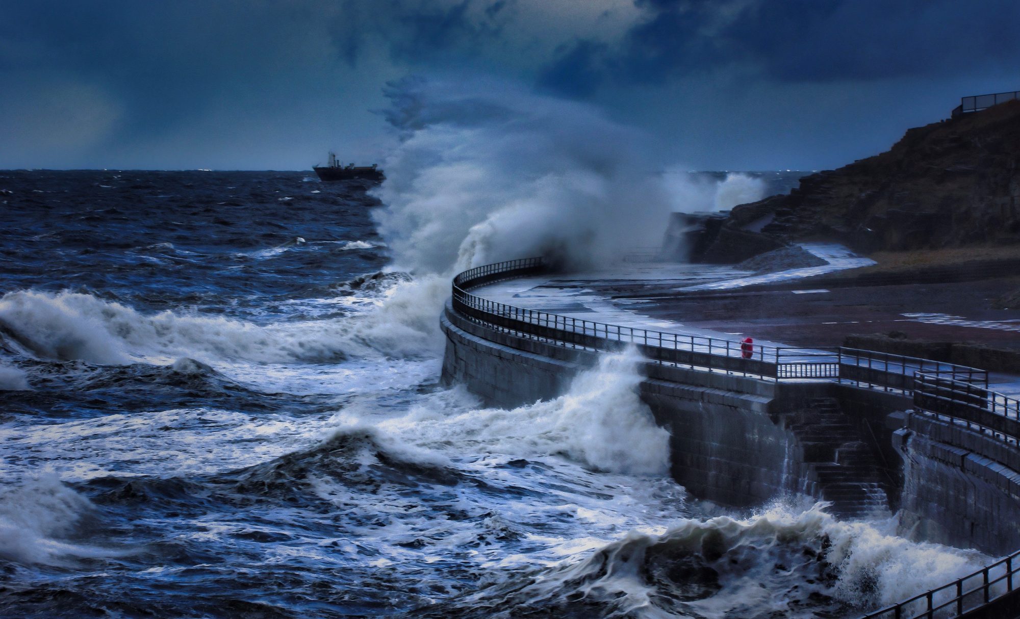 Large waves crash over a coastal seawall and walkway during a storm, illustrating the kind of elevated wave and flood risk facing low-lying coastlines.