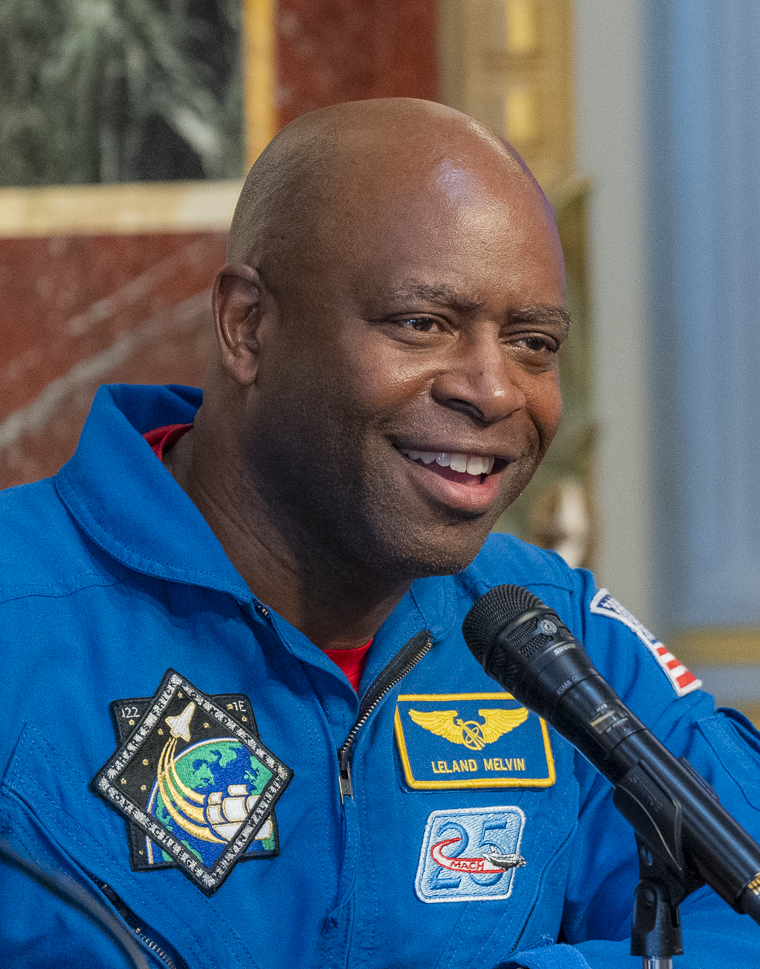 Man in blue NASA flight suit with name tag speaks at a microphone indoors.