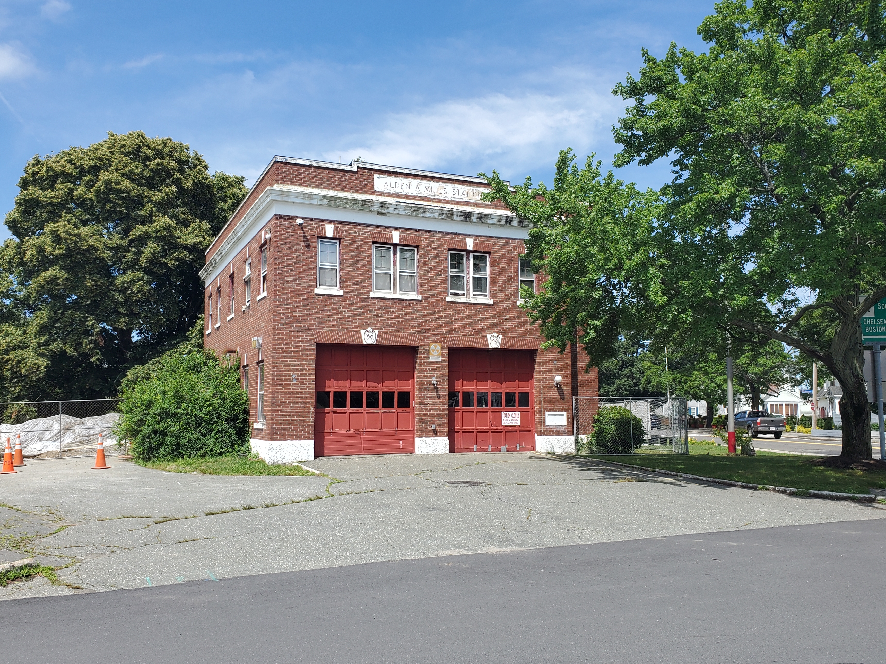 Two-story red brick fire station with two red garage bay doors, flanked by trees.