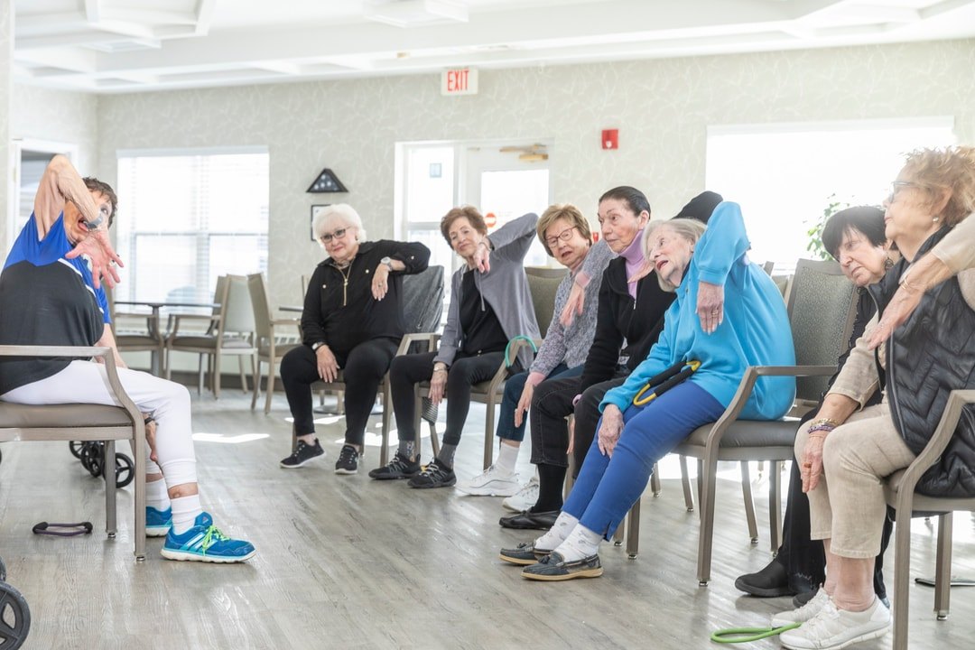 Elderly adults seated in chairs stretch to one side during a group exercise class.