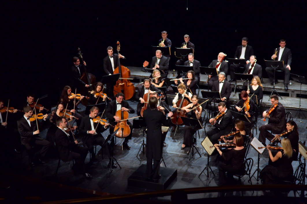 Symphony orchestra performing on stage with conductor at podium, viewed from above.