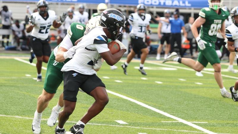 Football player in white uniform runs with the ball pursued by defenders in green.