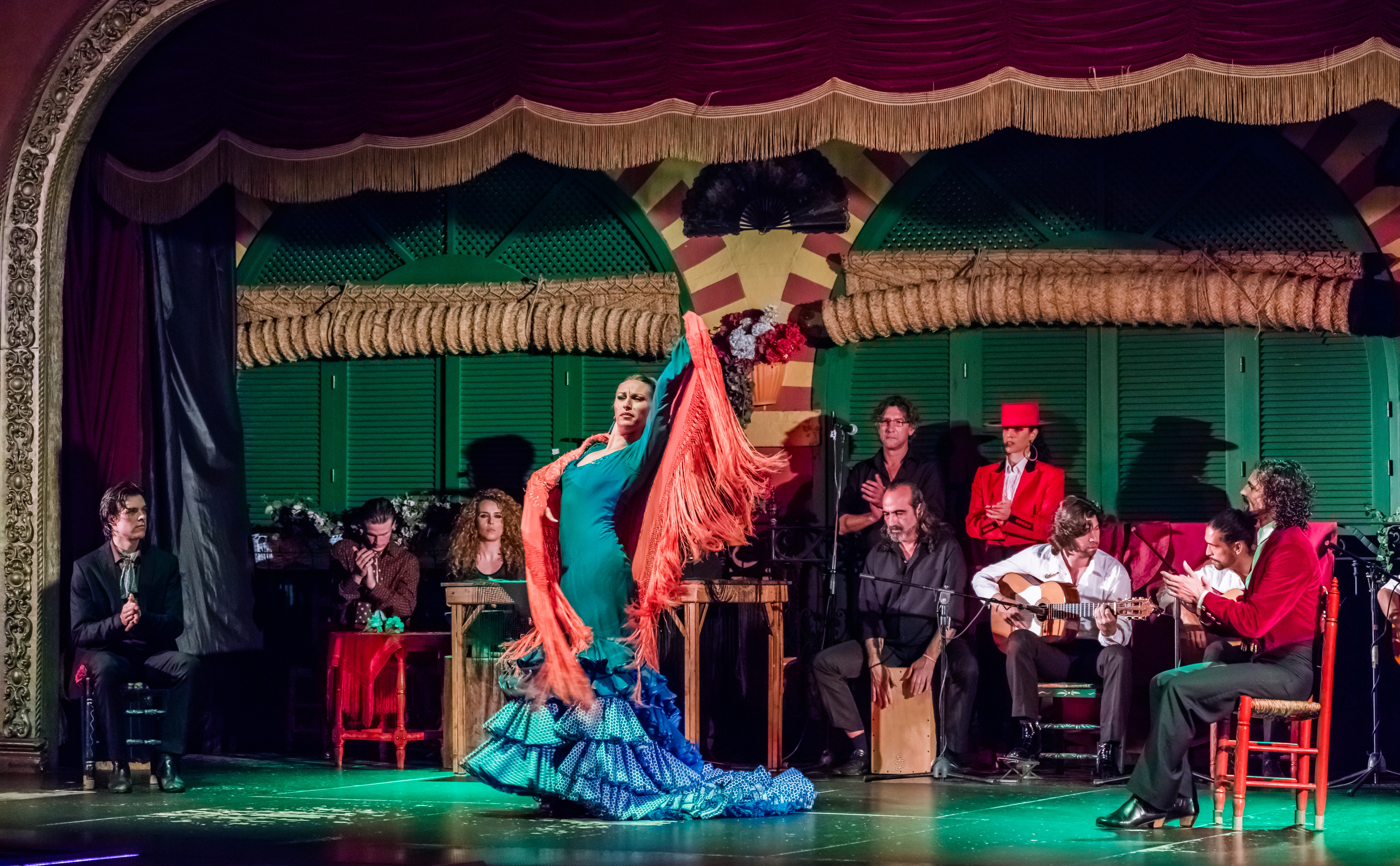 Flamenco dancer in teal ruffled dress on stage with seated guitarist and musicians behind her.
