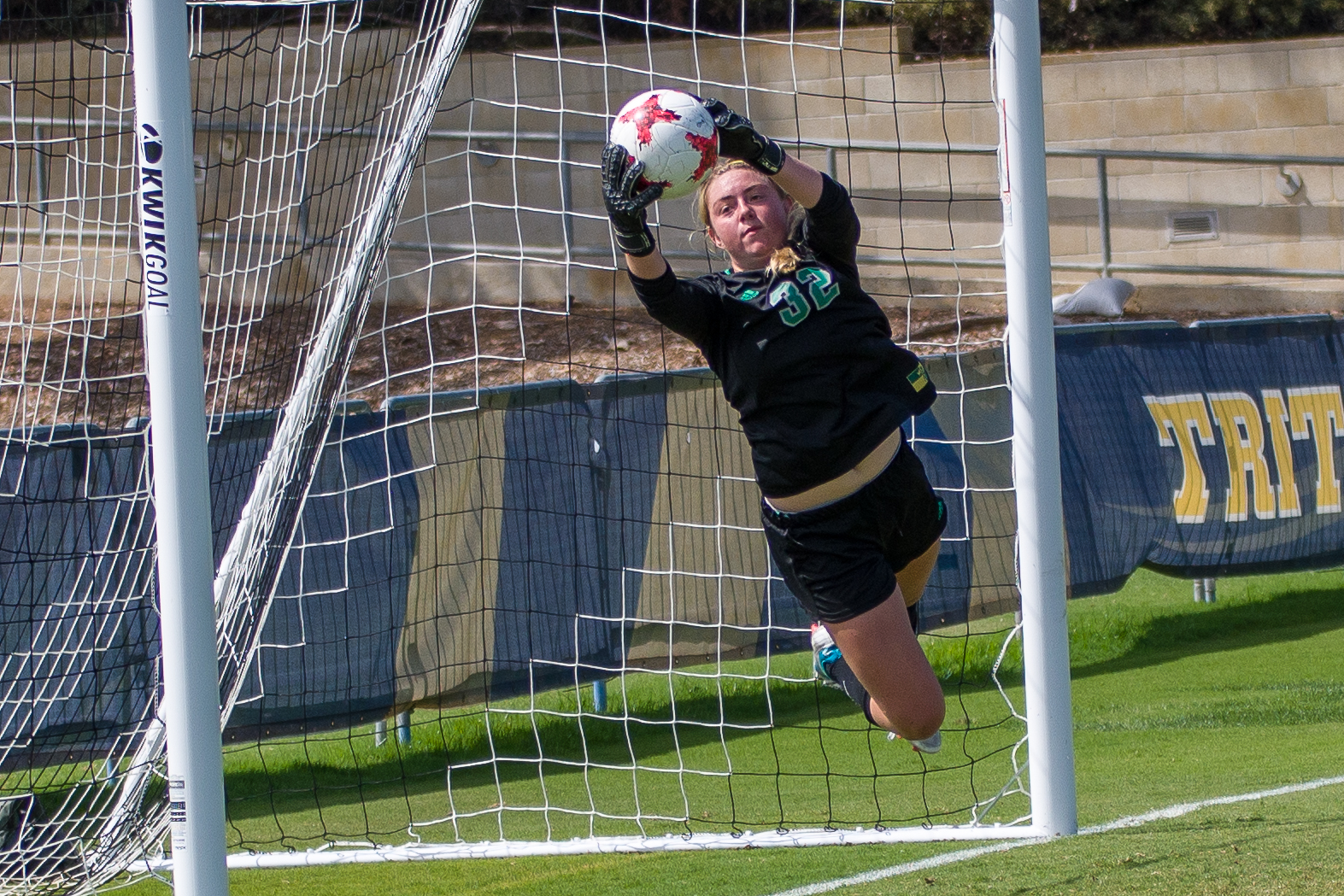 A goalkeeper in a dark uniform dives to catch a soccer ball in front of a goal net.