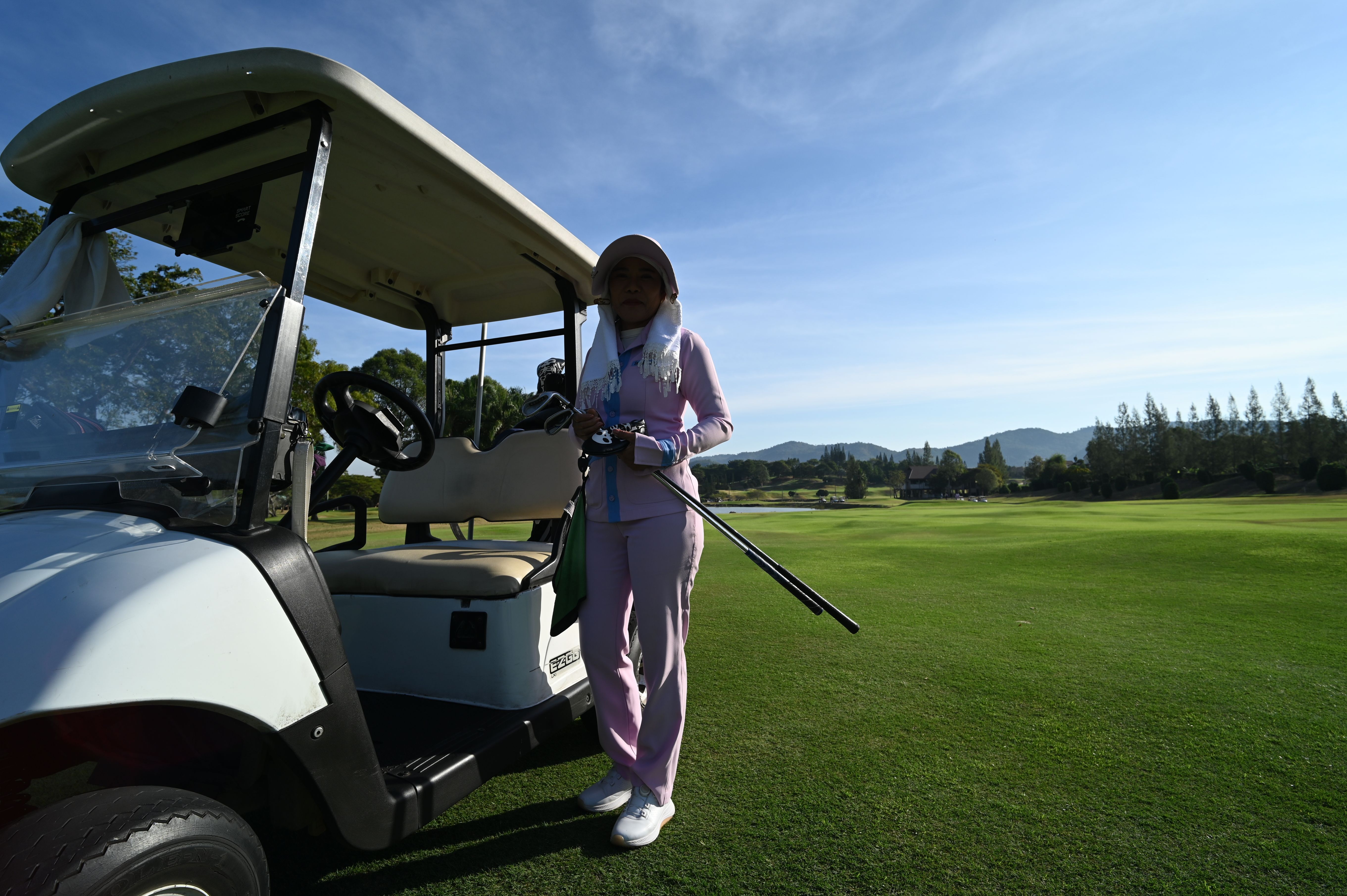 A golfer in a purple outfit stands next to a white golf cart on a green fairway.