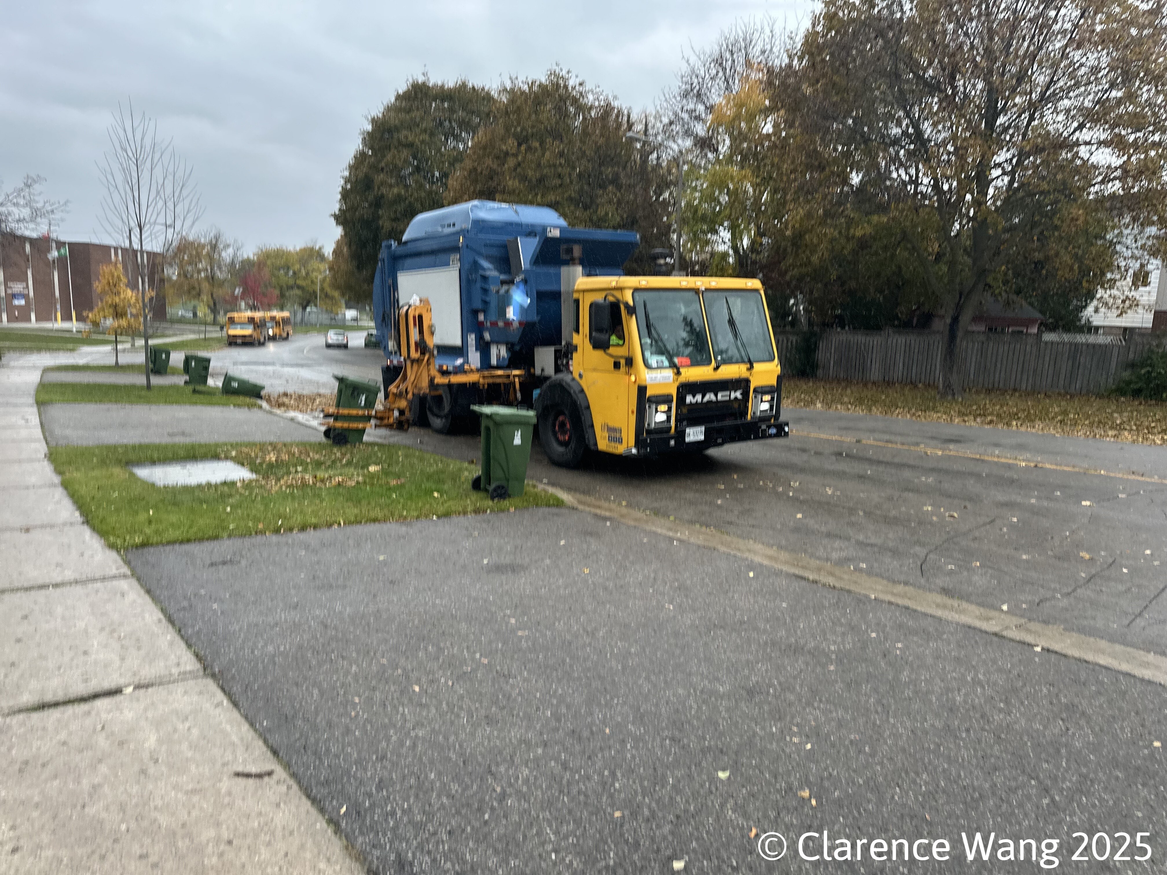 A yellow garbage truck collecting green bins on a residential suburban street.